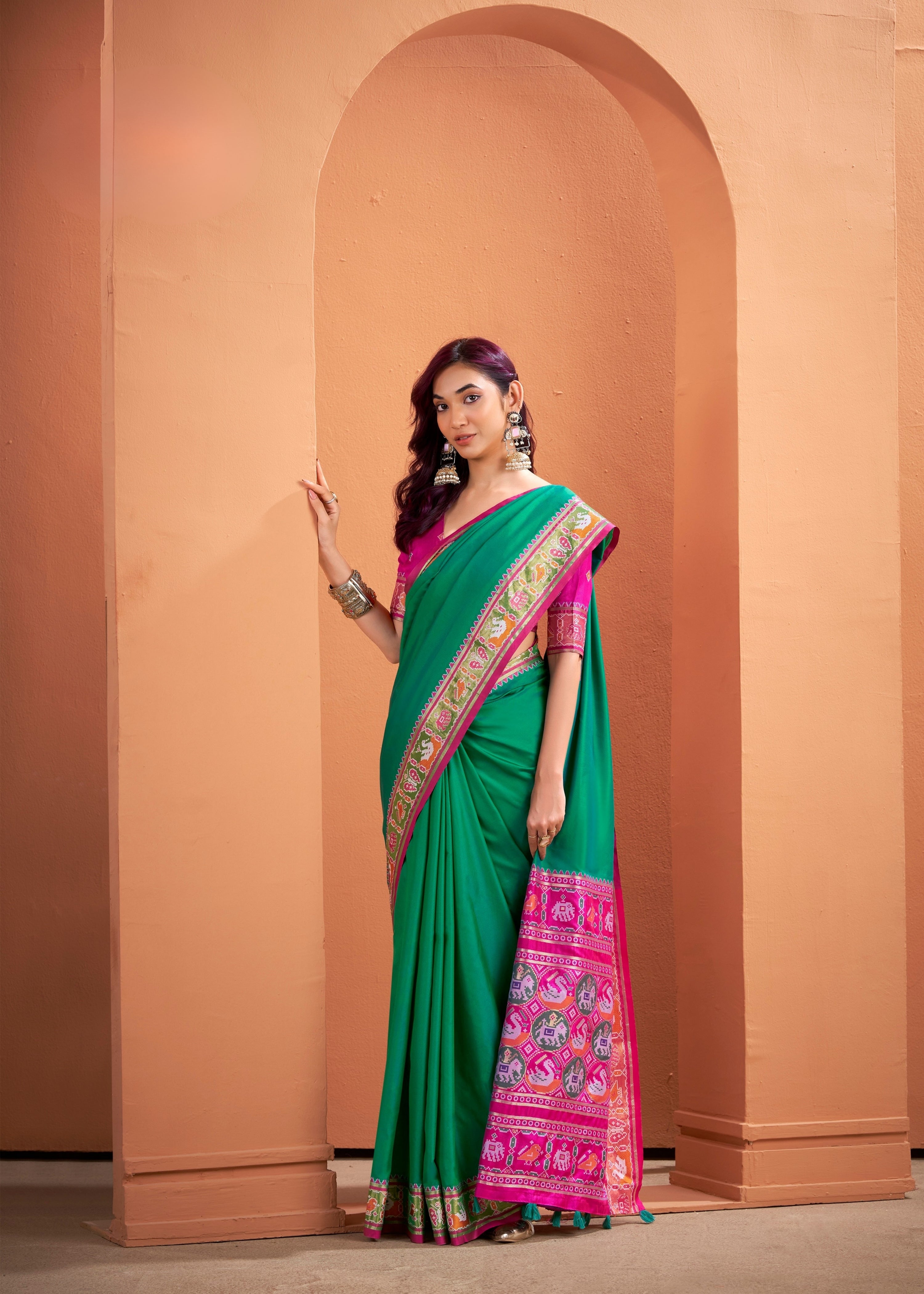 Woman wearing a green saree with pink border against a peach-colored wall.