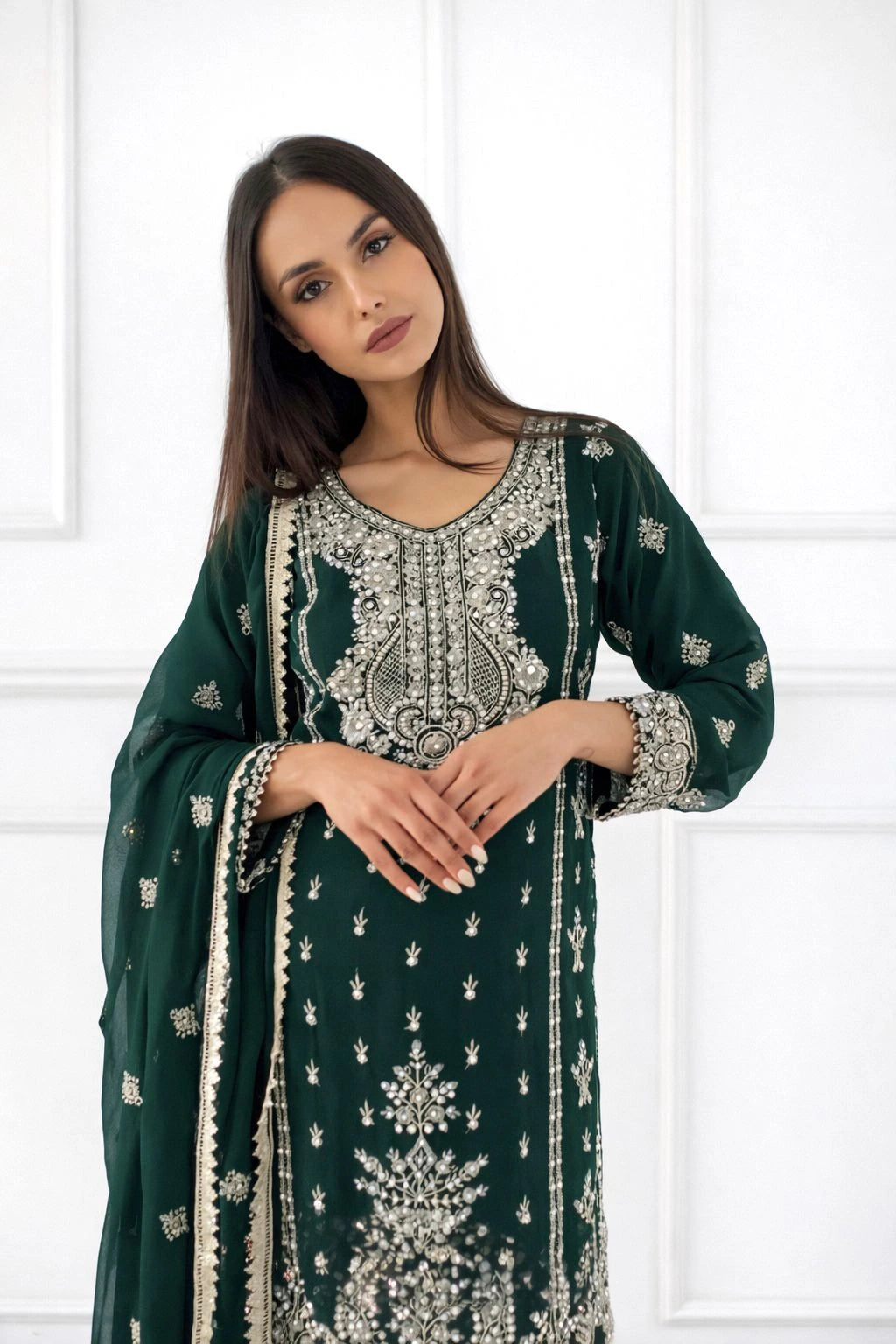 Woman wearing a green traditional outfit with intricate white embroidery against a white paneled wall.