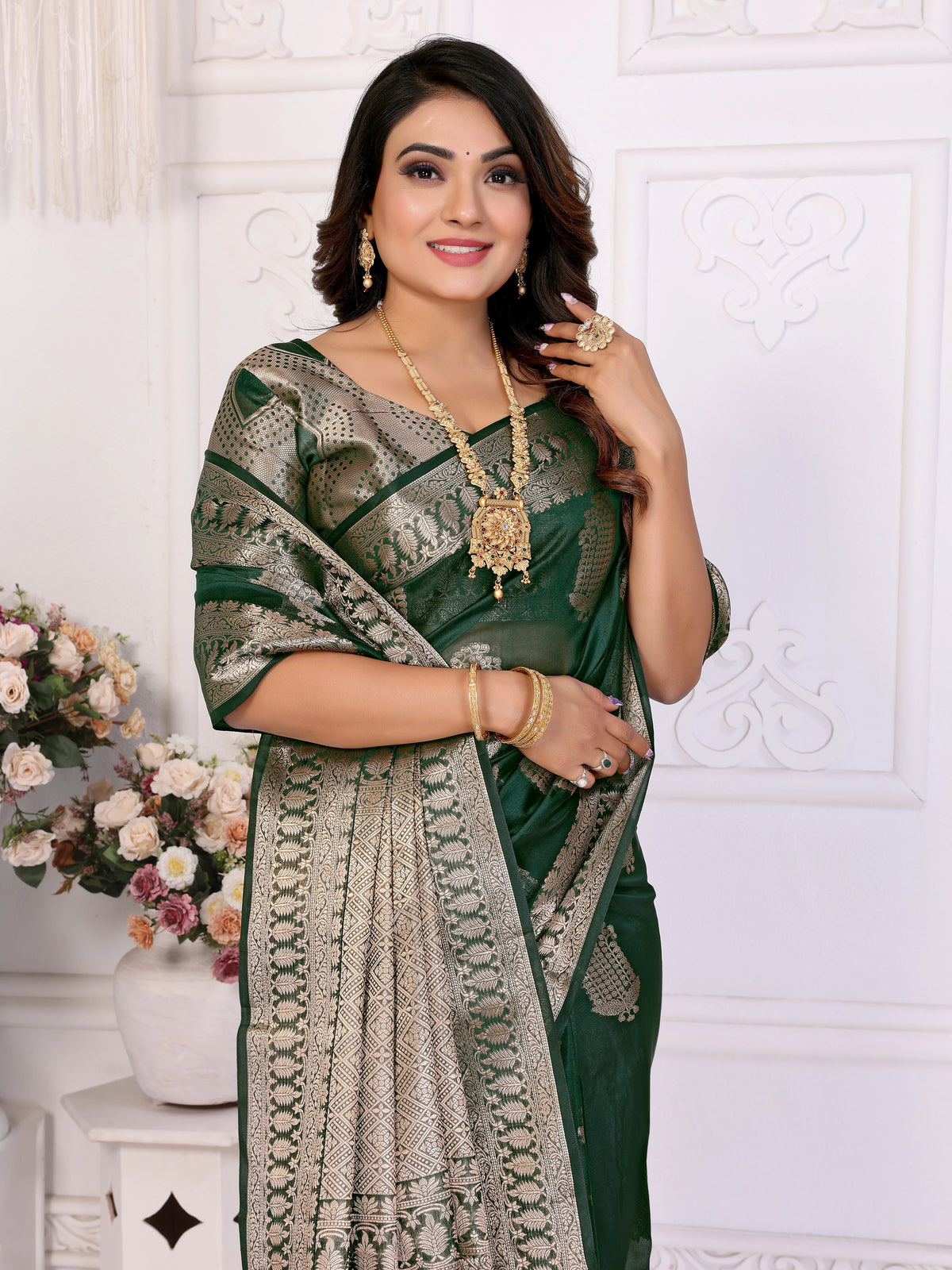 Woman in a green saree with gold patterns against a white decorative wall.