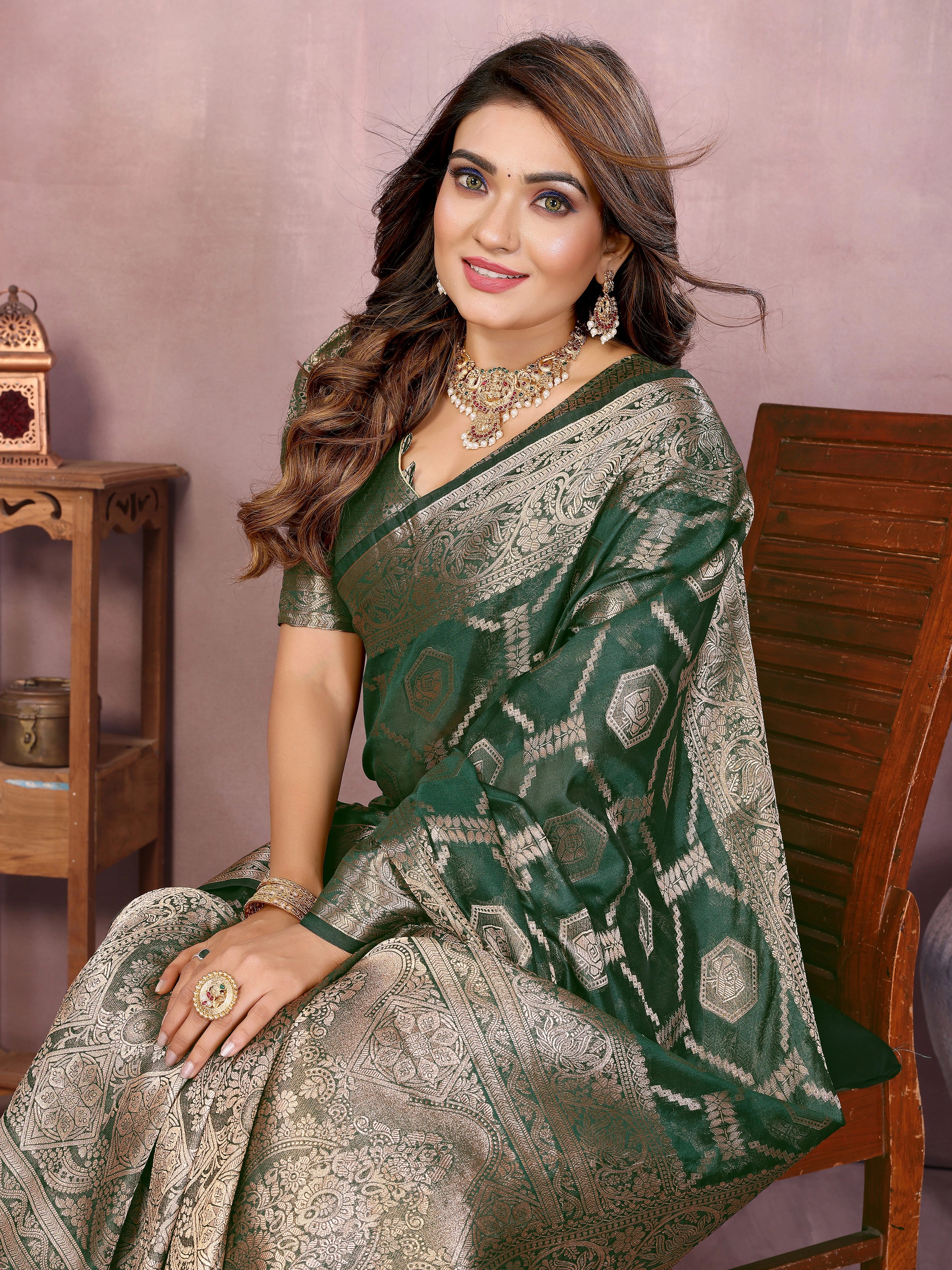 Woman in a green and silver saree sitting on a wooden chair against a pink wall.