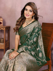 Woman in a green and silver saree sitting on a wooden chair against a pink wall.