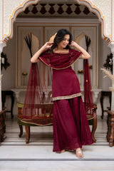 Woman in a burgundy saree with gold border standing in an ornate room.