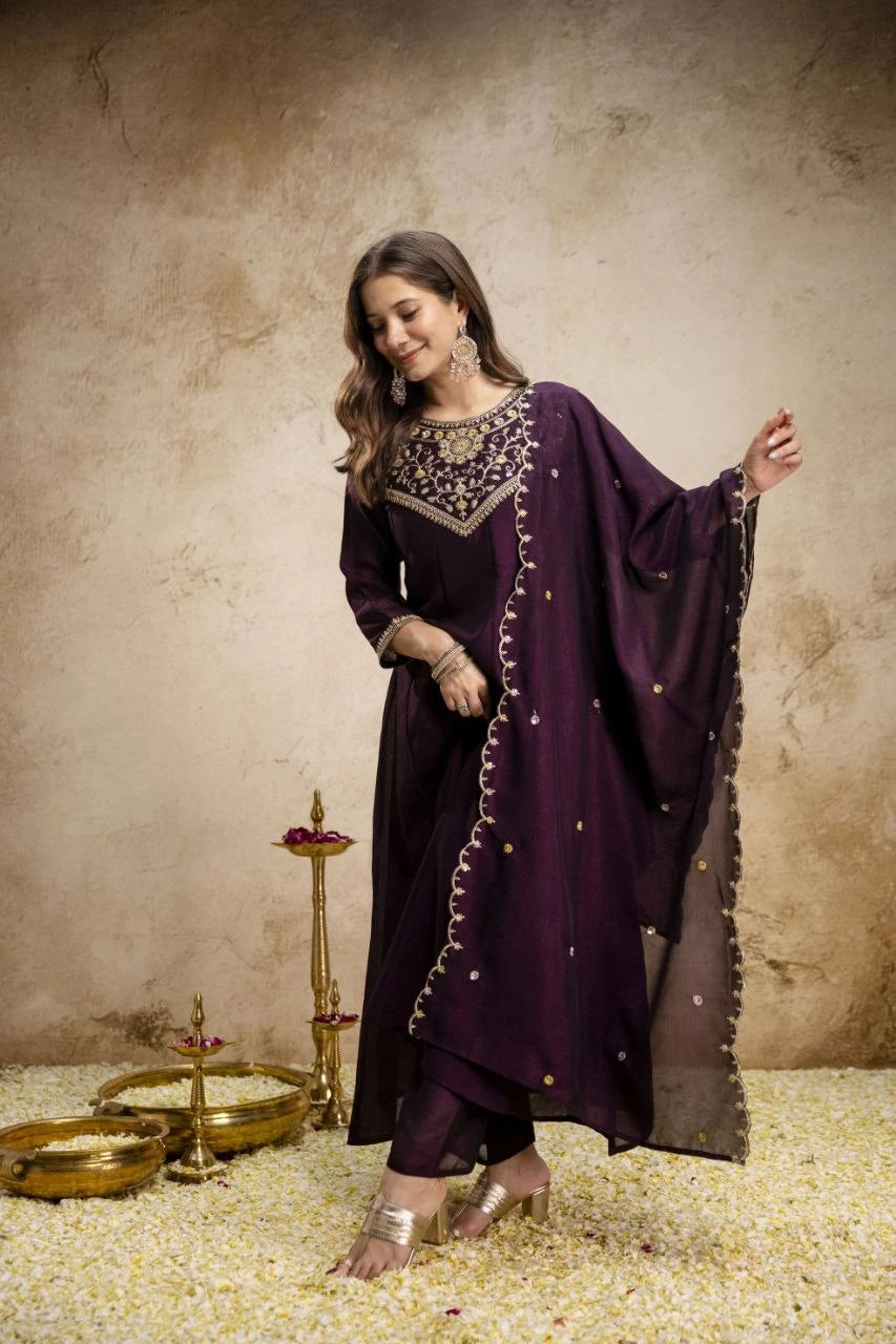 Woman in a dark purple traditional outfit with gold embellishments against a textured beige wall.