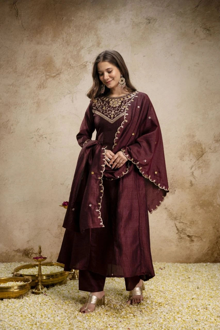 Woman in a maroon traditional outfit with gold embroidery against a textured beige wall.