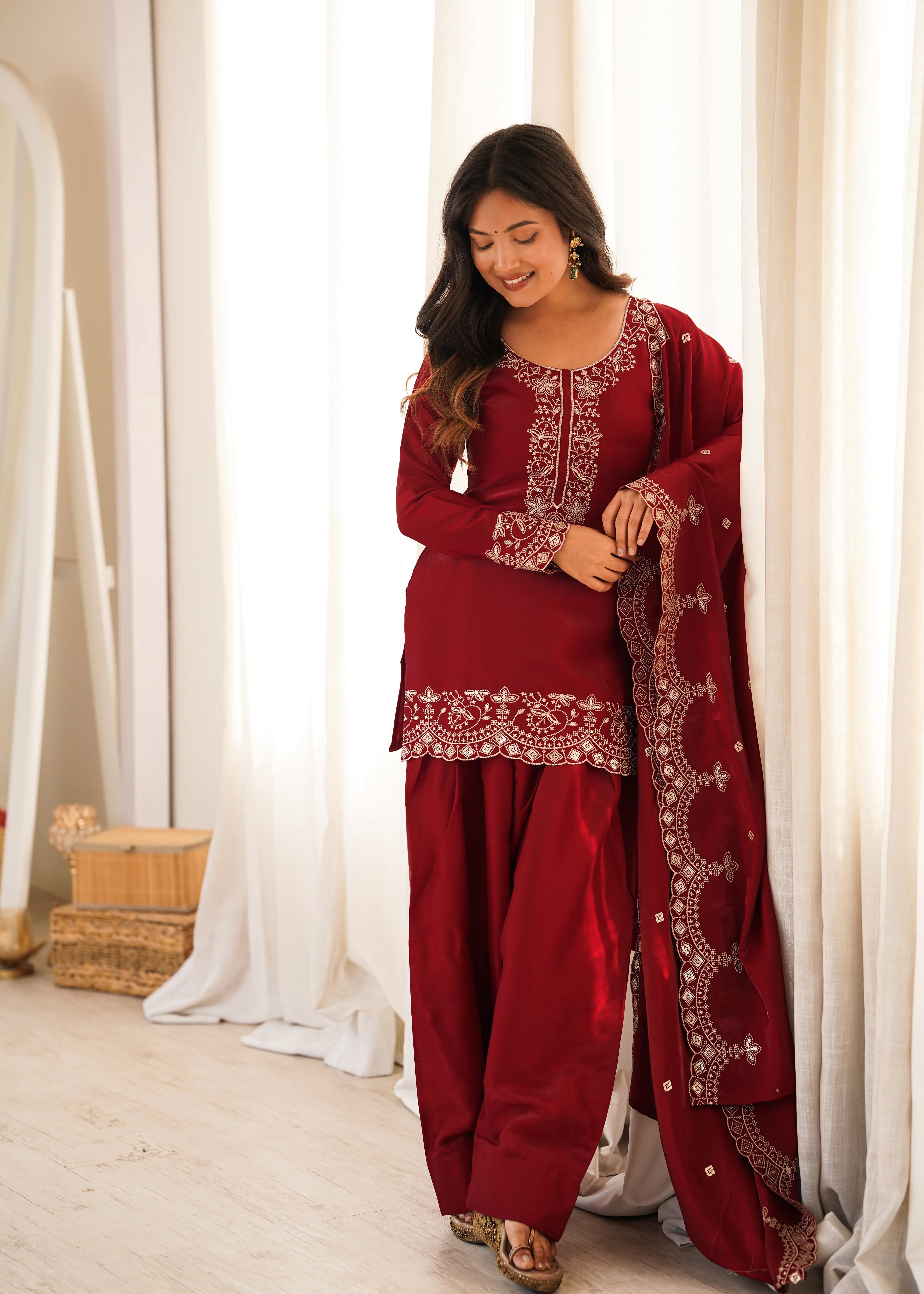 Woman in a red traditional outfit with white embroidery standing in front of white curtains.