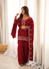Woman wearing a red embroidered traditional outfit in a bright room with white curtains.