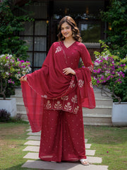 Woman in a red traditional outfit standing outdoors with greenery and flowers in the background