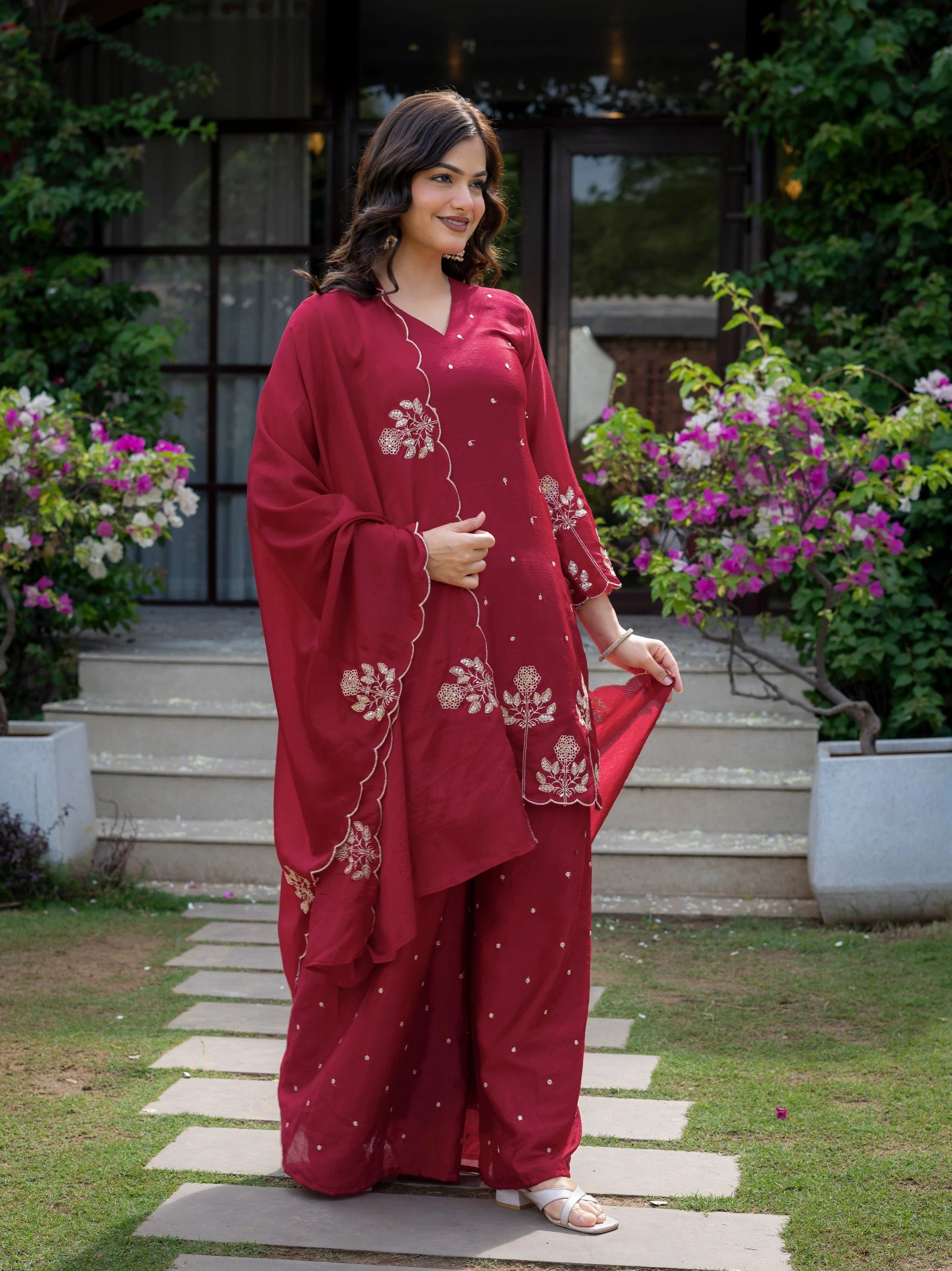 Woman in a red traditional outfit standing outdoors with flowers and steps in the background