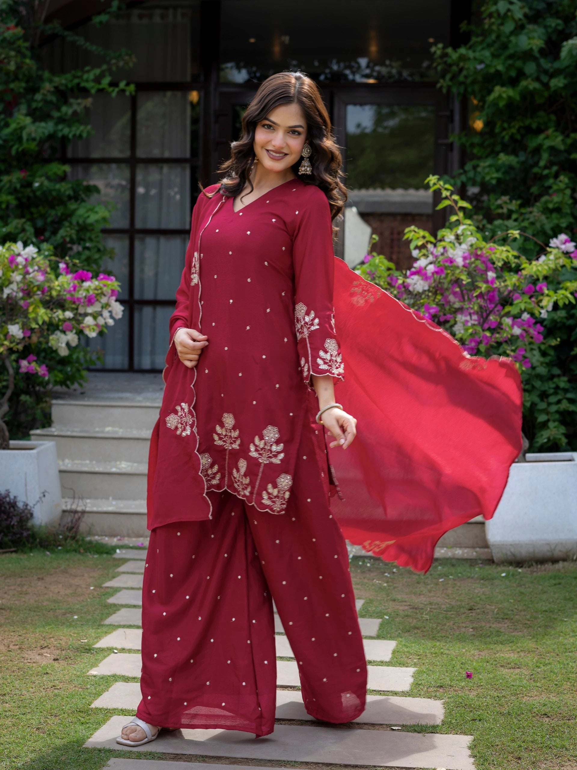 Woman in a red traditional outfit standing outdoors with flowers and greenery around.