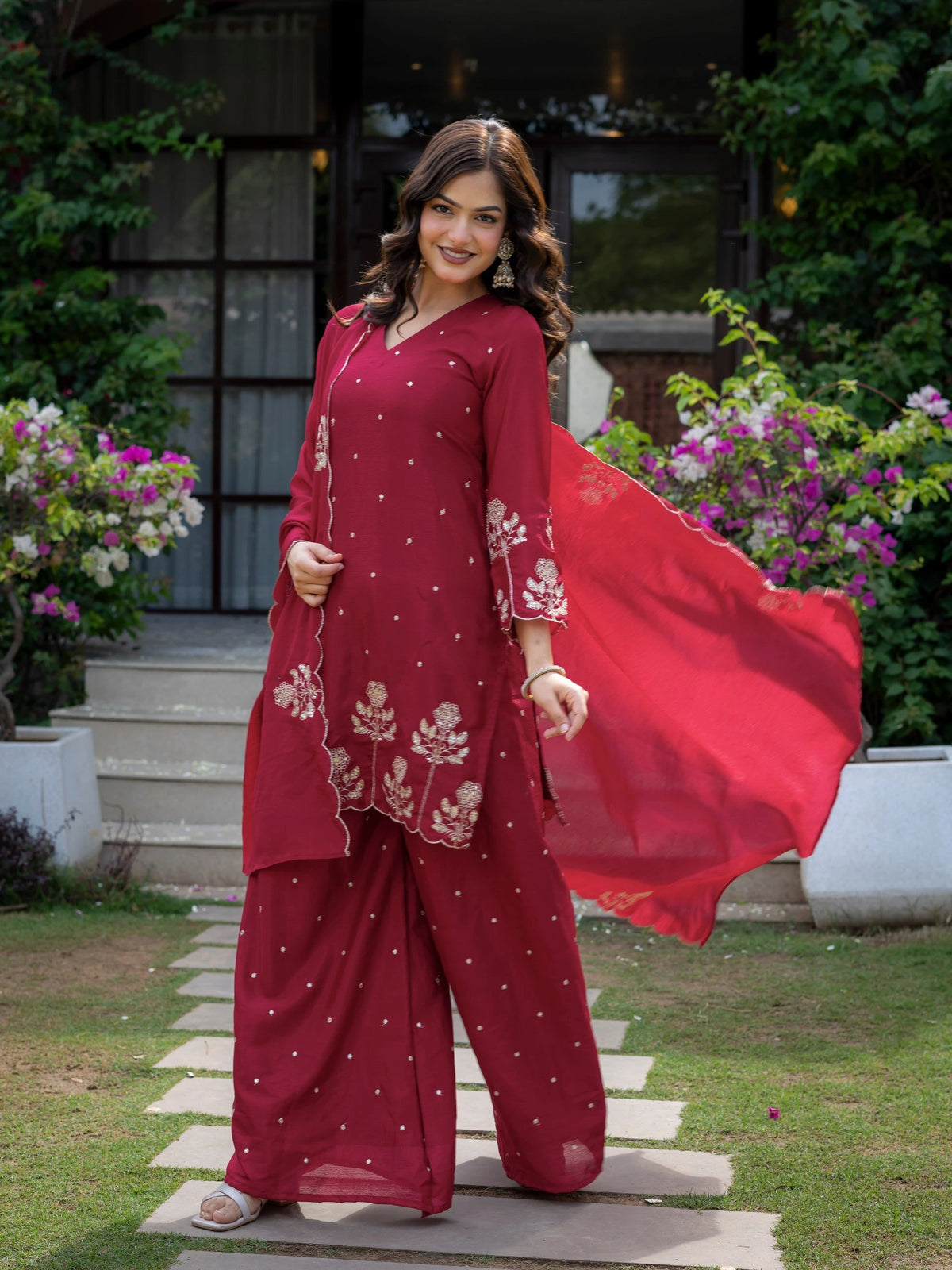 Woman in a red traditional outfit standing outdoors with flowers and greenery around.