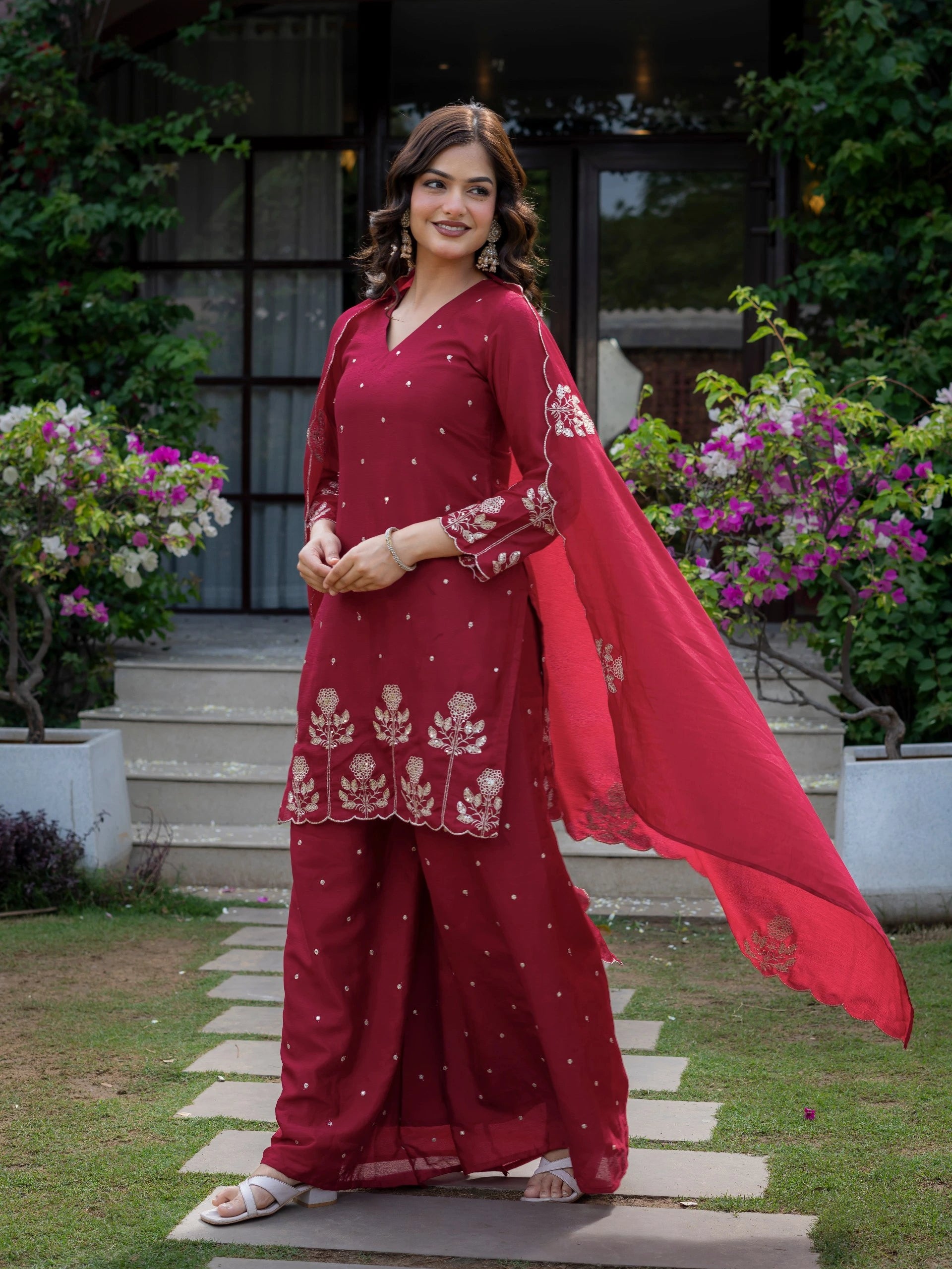 Woman in a red traditional outfit standing outdoors with flowers and a building in the background