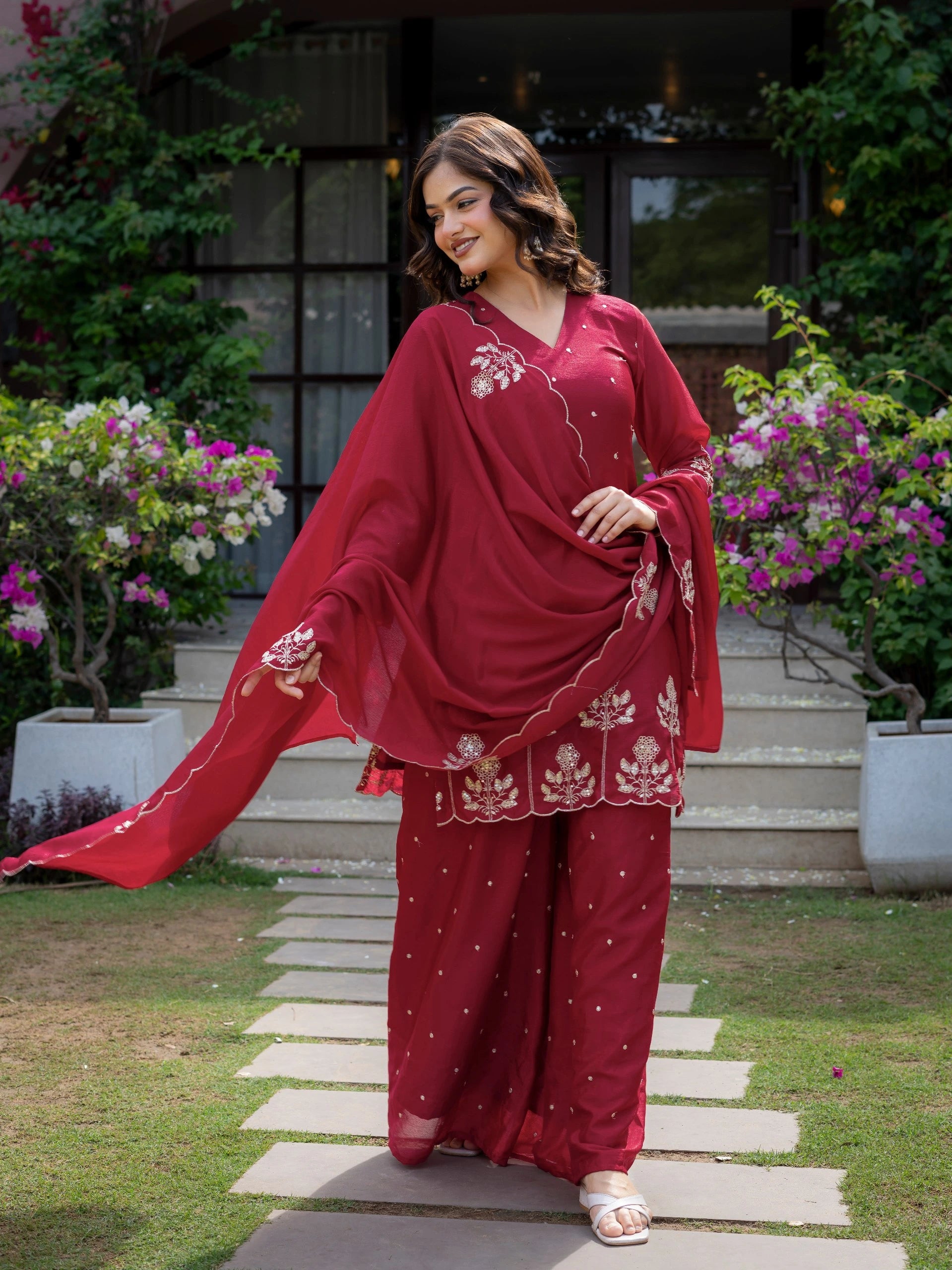 Woman in a red traditional outfit with floral patterns standing on a stone path with flowers and plants in the background.