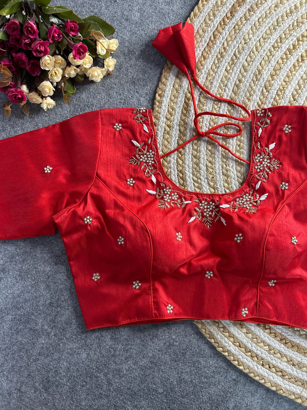 The front of the red raw silk blouse showcases a curved V-neckline decorated with the same intricate white and gold floral embroidery. Small, scattered floral buttis are embroidered across the bodice and sleeves, and the blouse is laid flat next to a bouquet of pink and white roses on a textured grey background.
