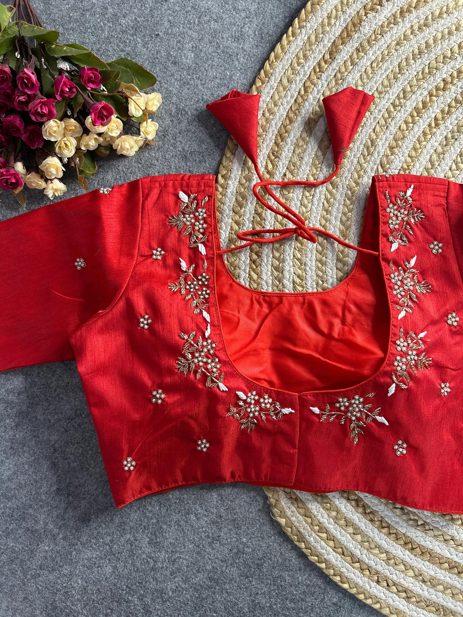 A vibrant red raw silk blouse featuring a deep U-shaped back. The neckline is adorned with delicate hand-embroidered floral sprigs in white and gold thread. Matching red tie-back tassels are attached at the shoulders, and the rest of the back is sprinkled with small embroidered floral motifs.