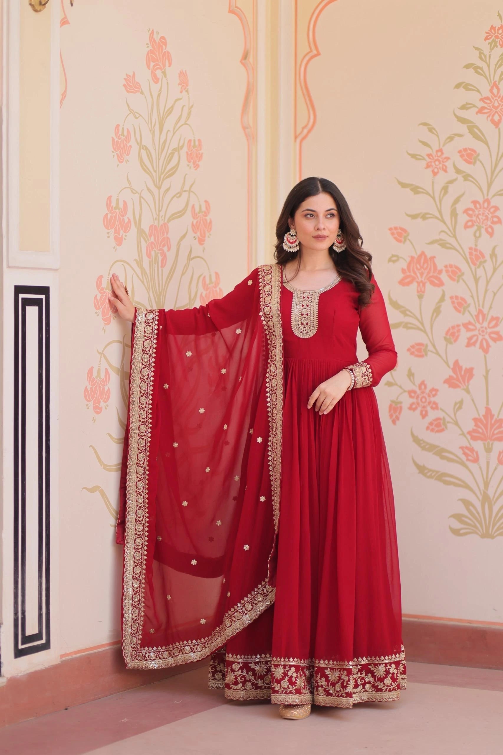 Woman in a red traditional outfit with gold embroidery against a decorative wall.