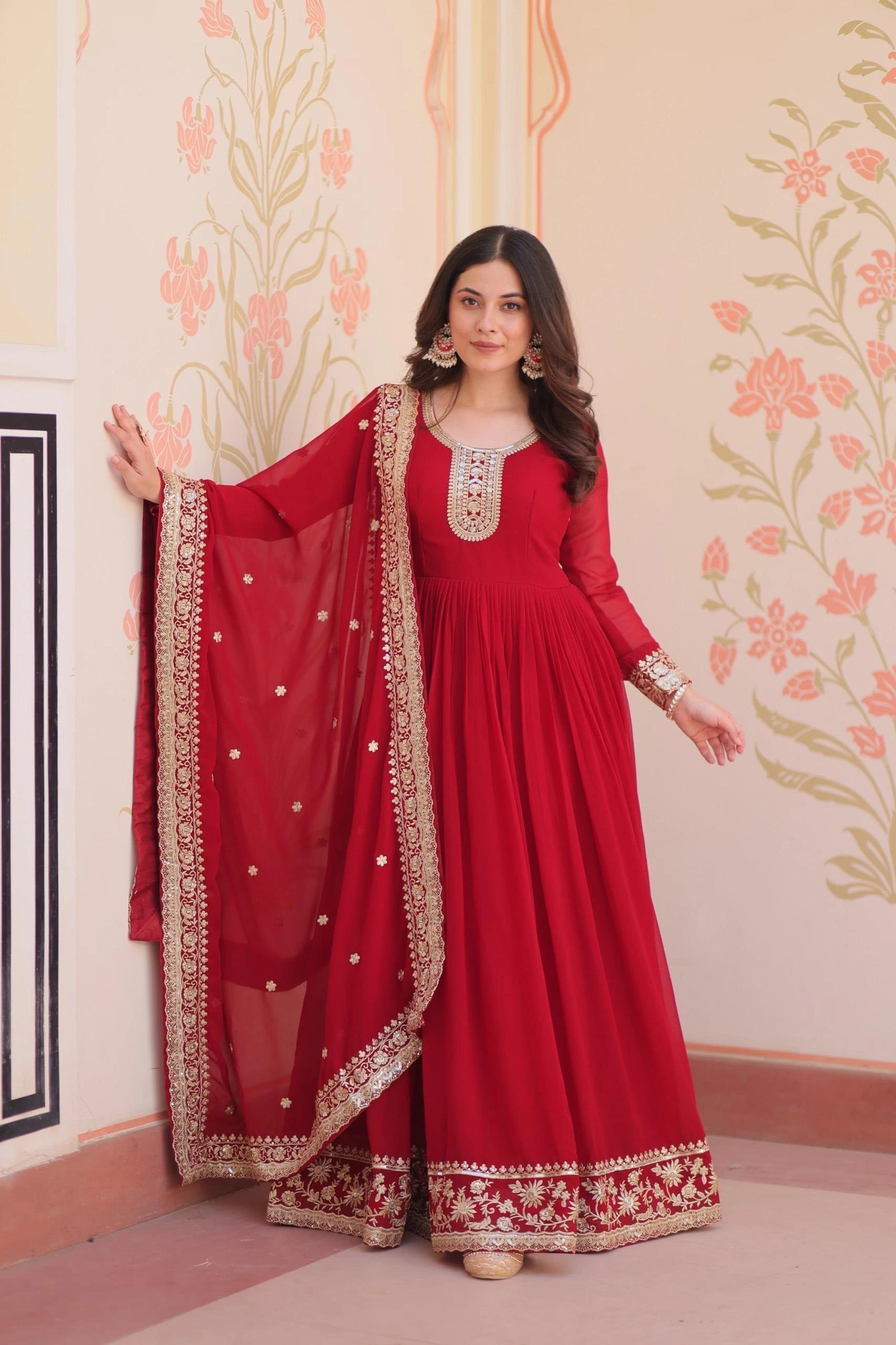 Woman in a red traditional outfit with gold embroidery against a floral wall.