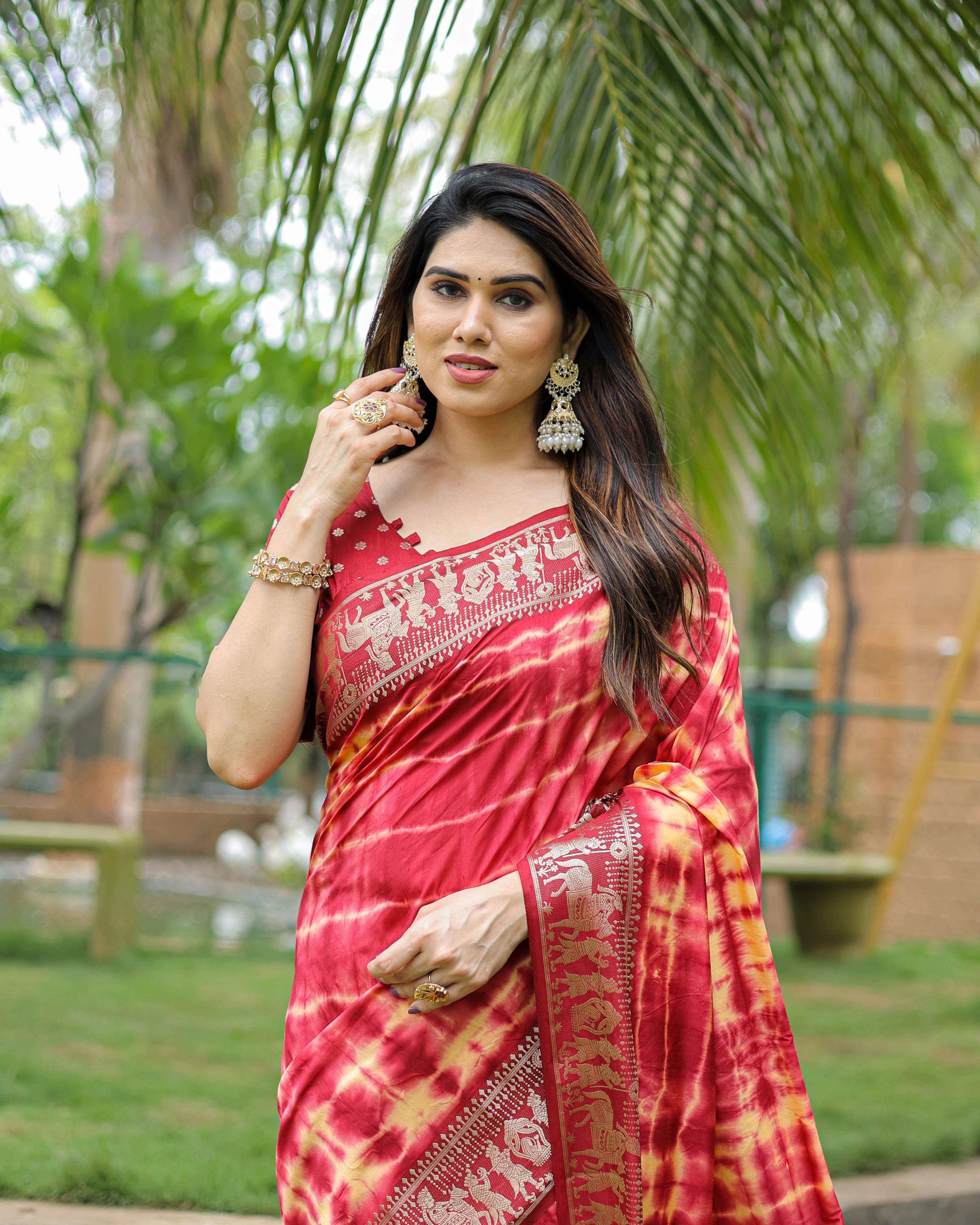 Woman in a red saree with white patterns standing outdoors with greenery in the background