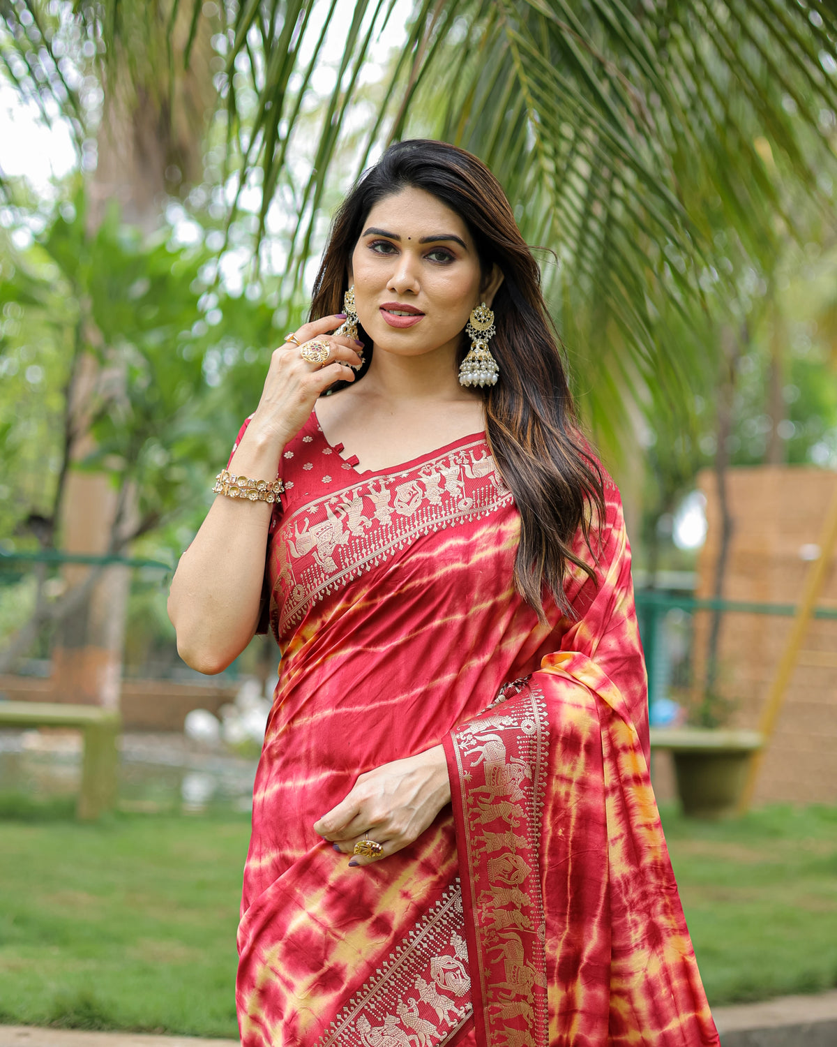 Woman in a red saree with white patterns standing outdoors with greenery in the background
