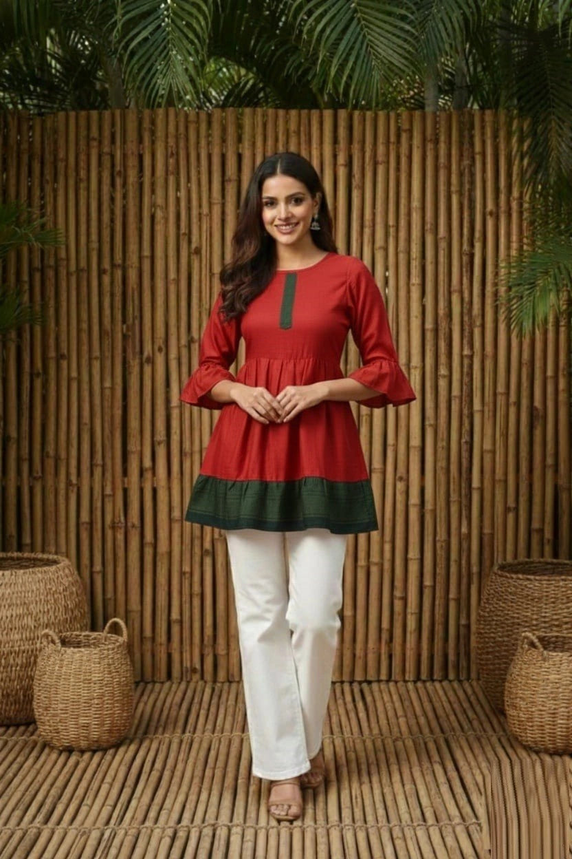 Woman wearing a red and green top with white pants standing against a wooden wall.