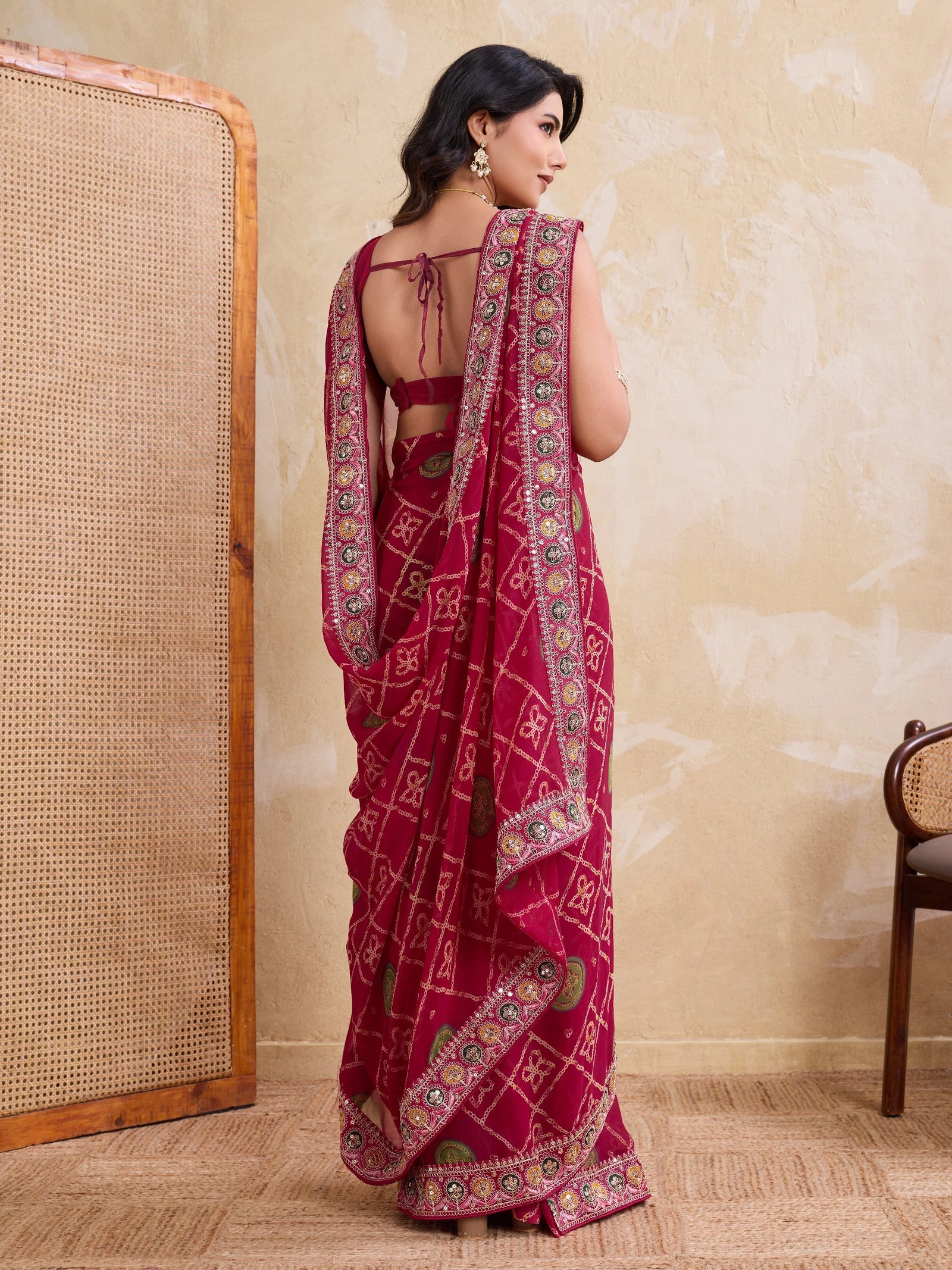 Woman wearing a red saree with intricate patterns in a room with a textured wall and wooden chair.
