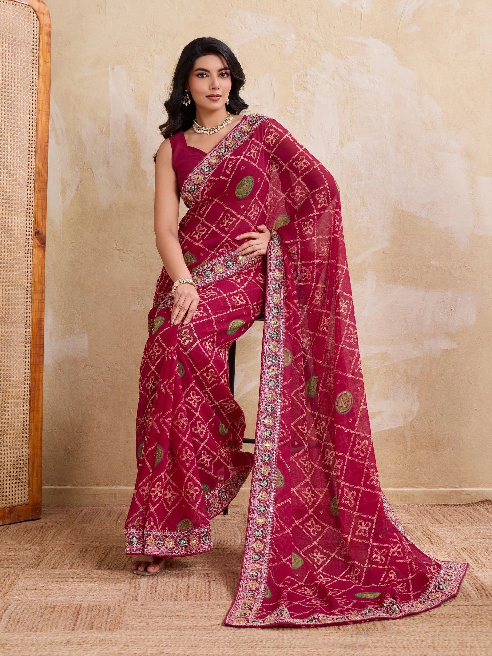Woman wearing a red saree with intricate patterns against a beige wall.