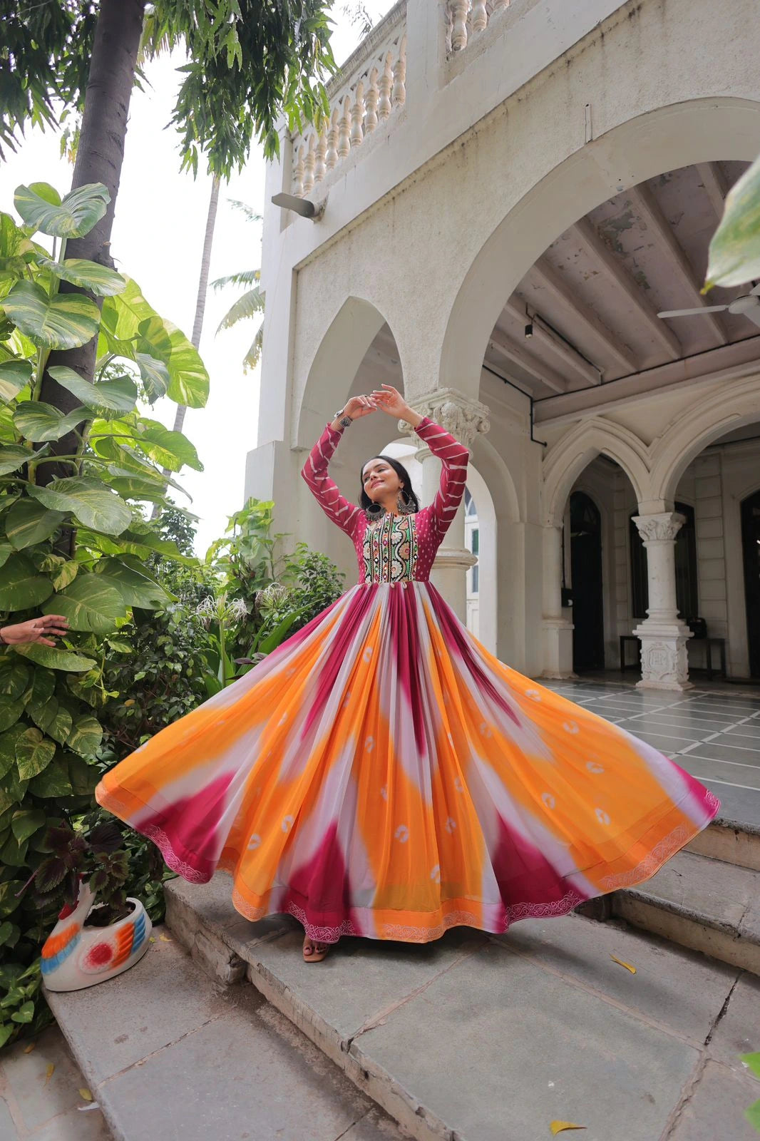 Woman in a colorful traditional dress standing on steps with a building and greenery in the background