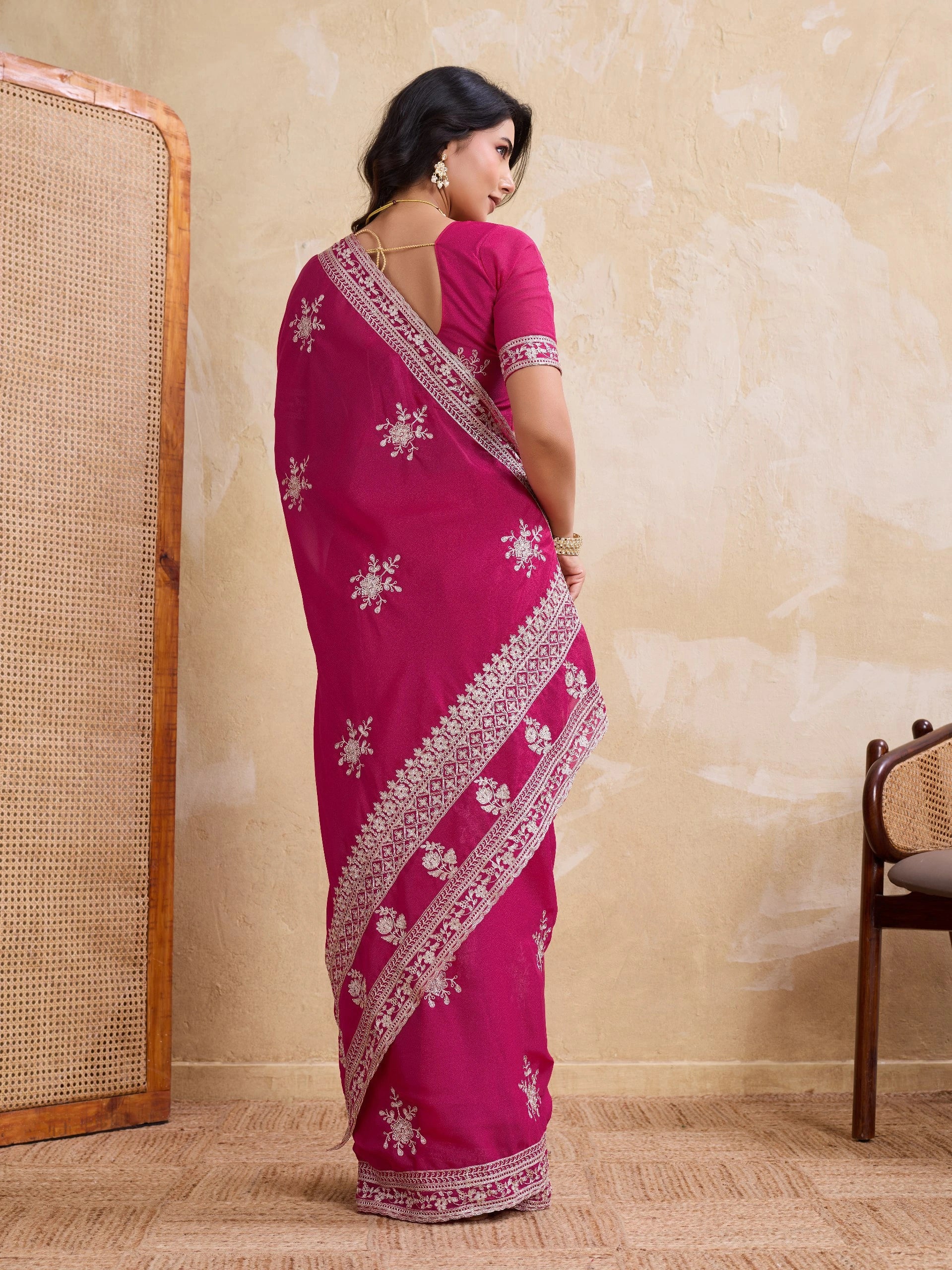 Woman wearing a pink saree with white patterns in a room with a beige wall and wooden chair.