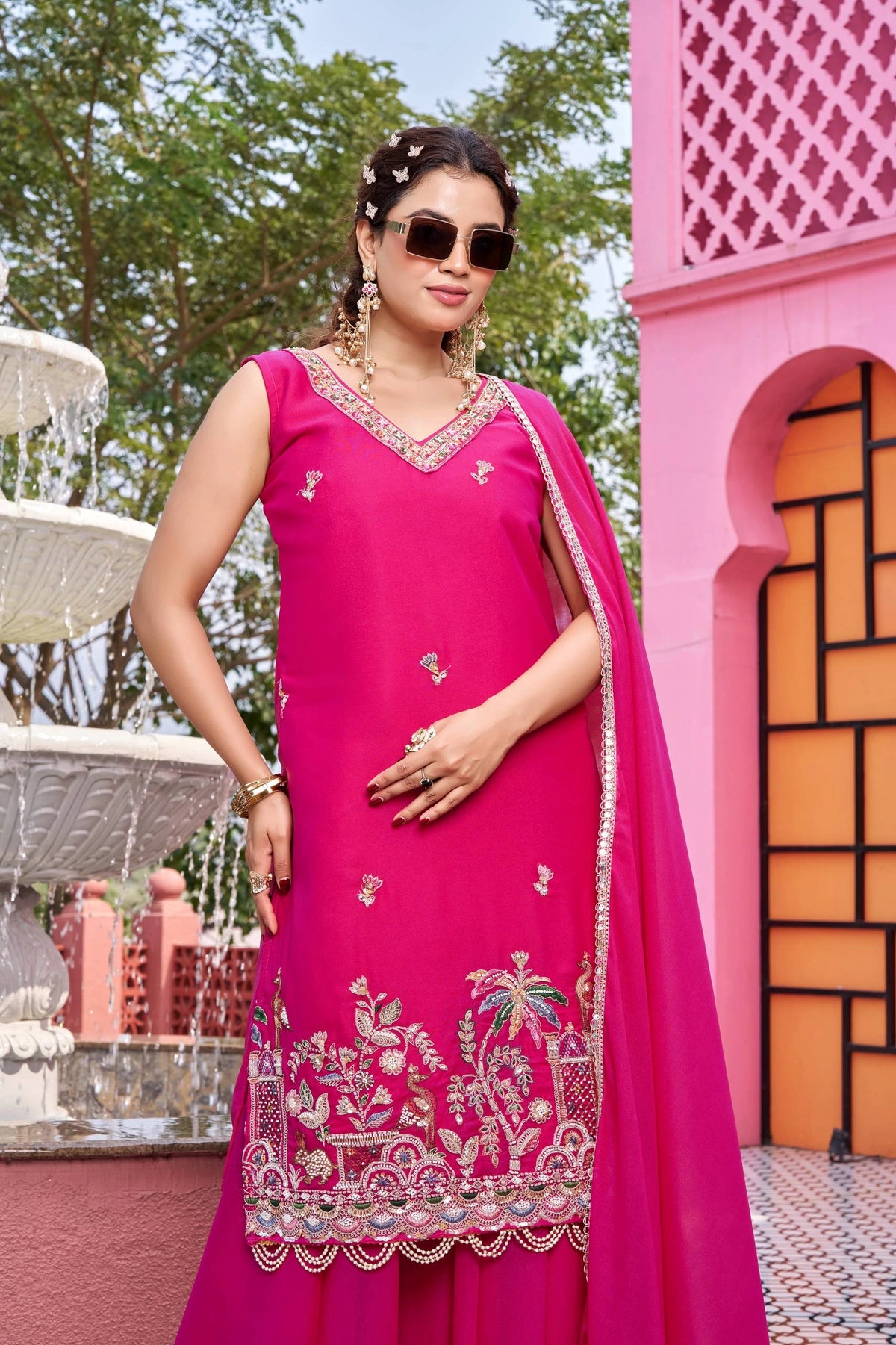 Woman in a pink traditional outfit with embroidery standing in front of a pink architectural structure.
