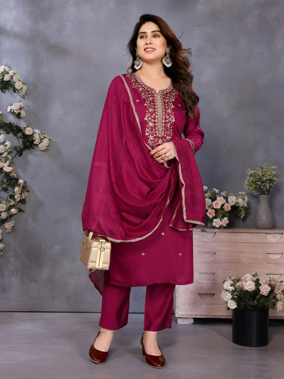 Woman in a magenta traditional outfit with embroidery, standing against a gray background with decorative elements.