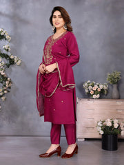 Woman in a magenta traditional outfit with gold embroidery standing against a gray background with decorative elements.