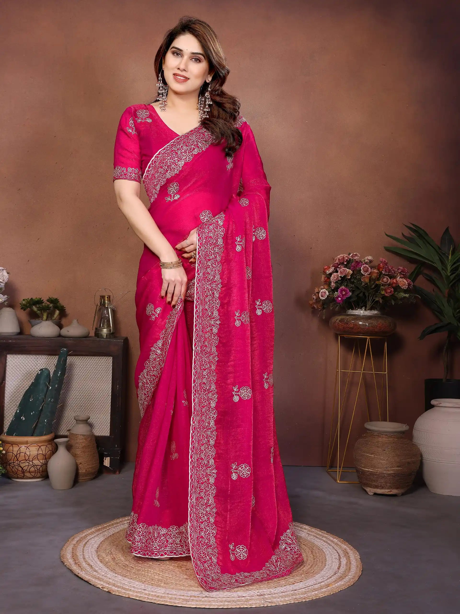 Woman in a bright pink saree with silver border standing in a room with decorative plants and vases.