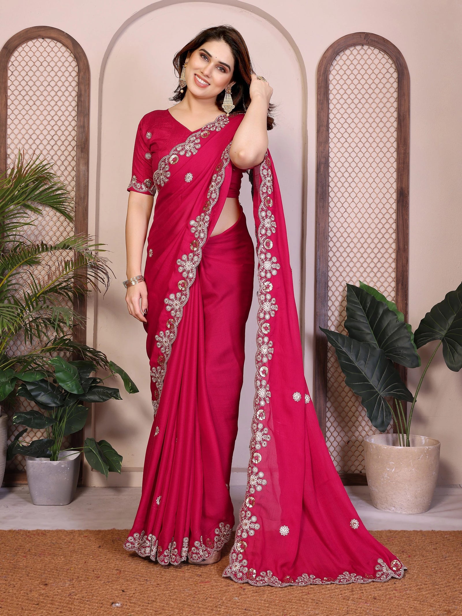 Woman wearing a red saree with white patterns in an indoor setting with plants.