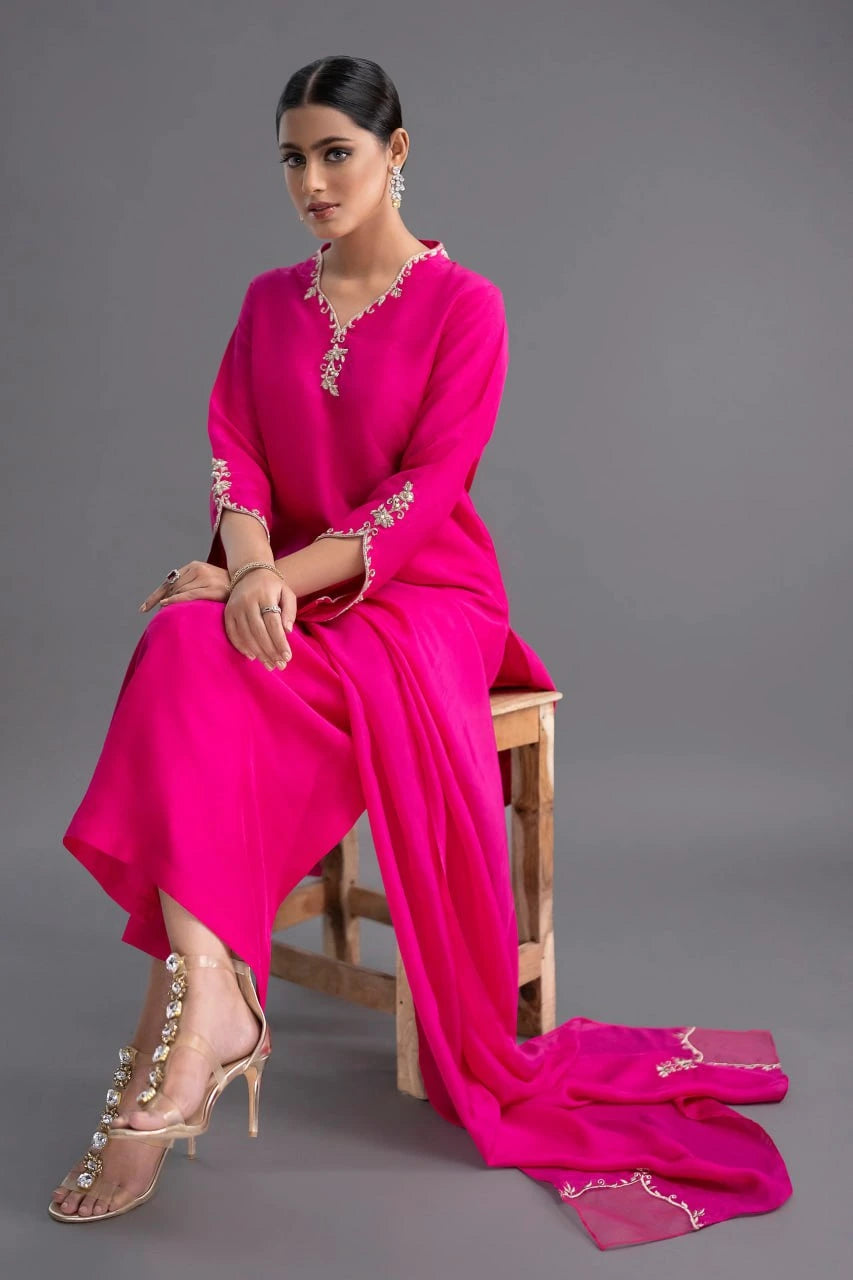 Woman in a bright pink traditional outfit with intricate designs, sitting on a wooden stool against a gray background.