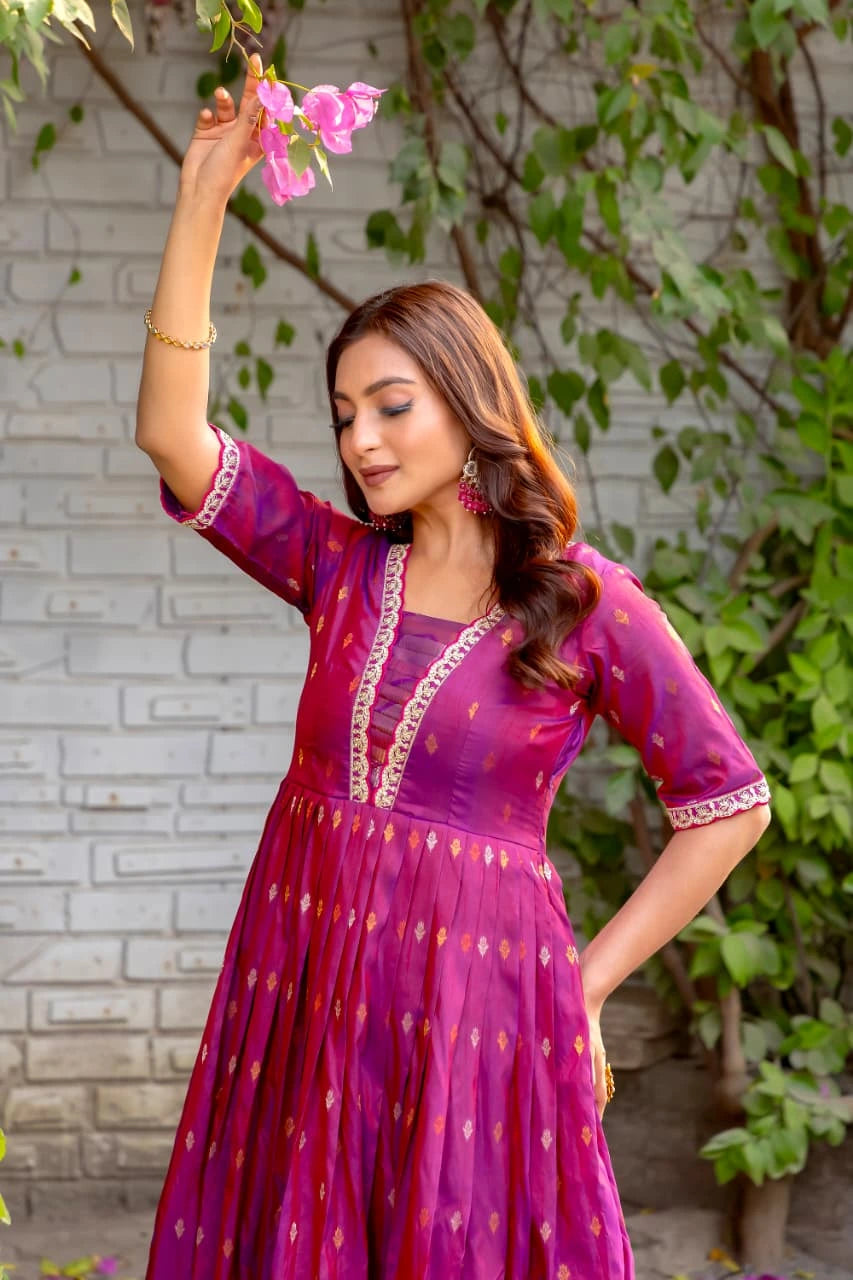 Woman in a purple traditional outfit holding pink flowers against a brick wall and greenery background