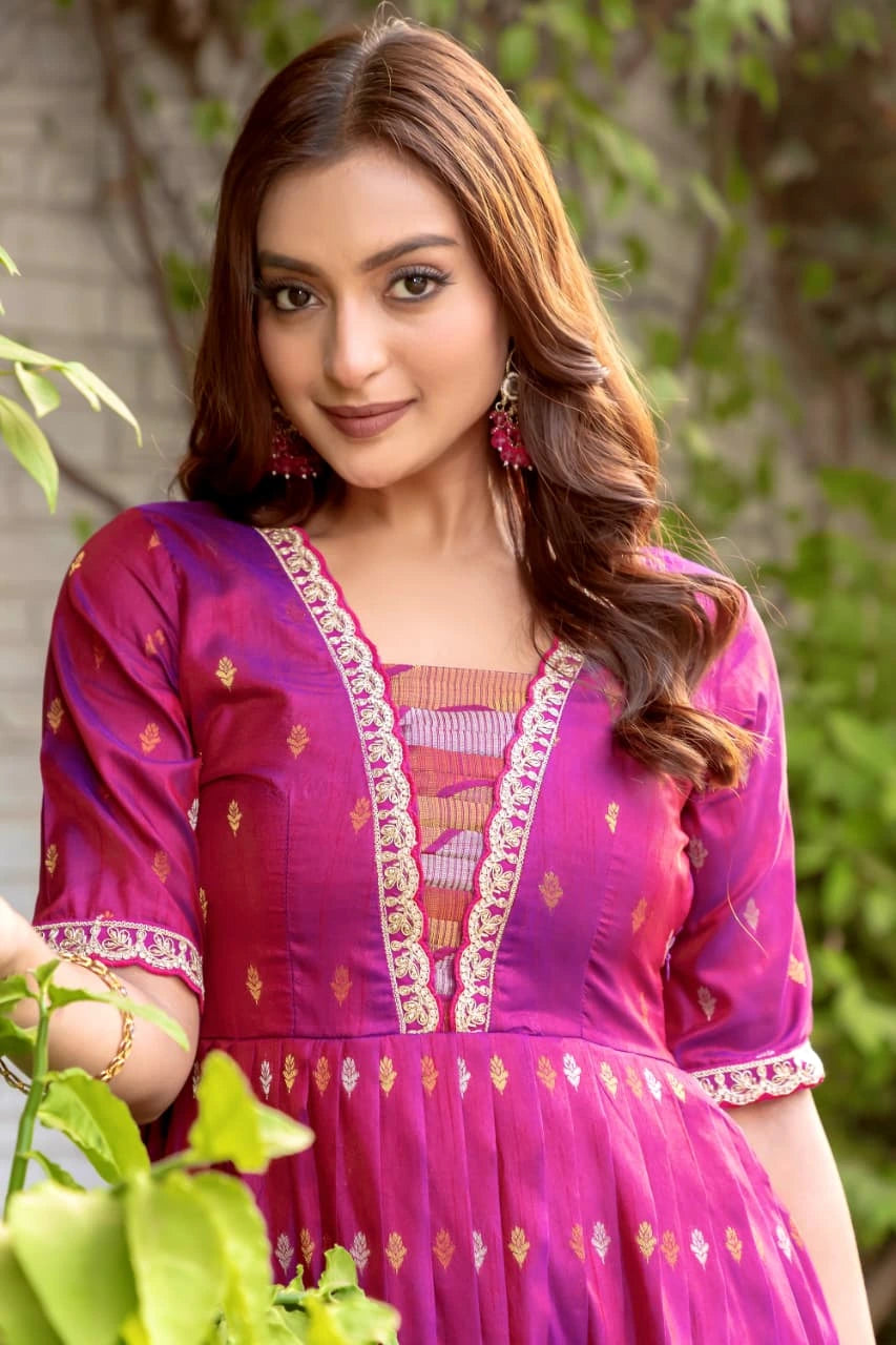 Woman wearing a pink traditional outfit with intricate designs, standing outdoors with greenery.
