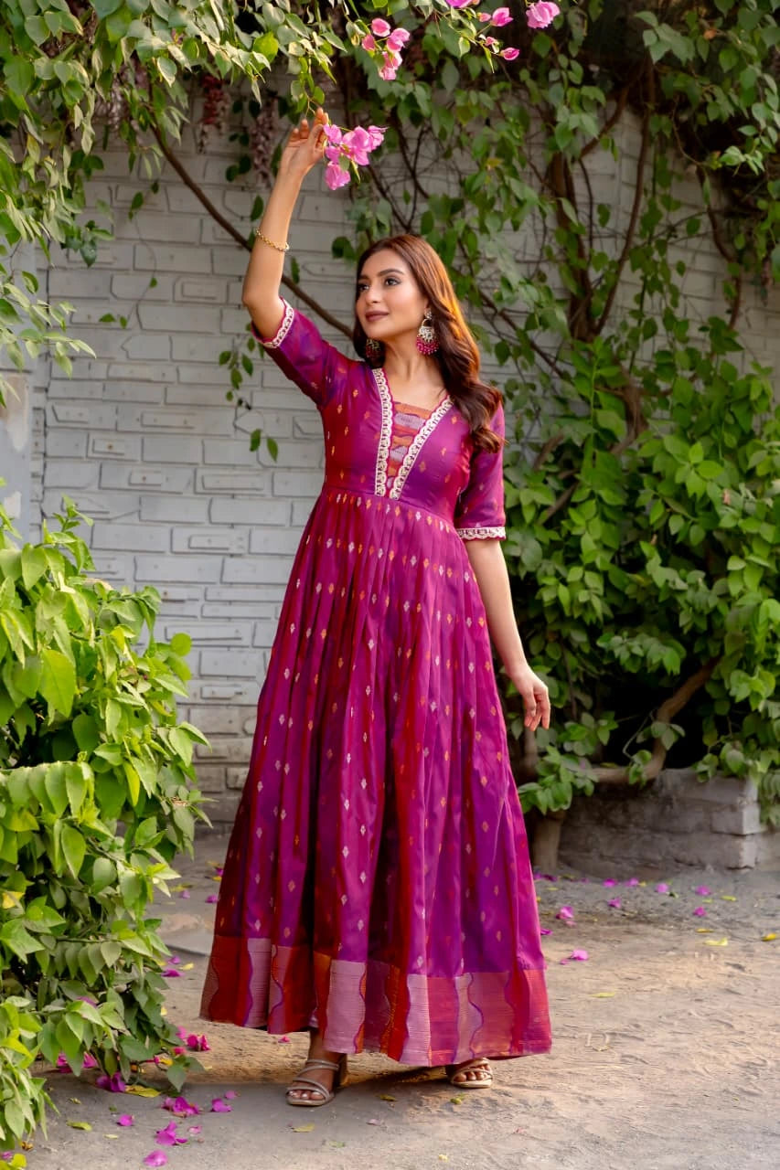 Woman in a purple dress standing outdoors with greenery and flowers around.