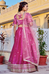 Woman in a pink traditional outfit standing in front of a yellow building with plants.