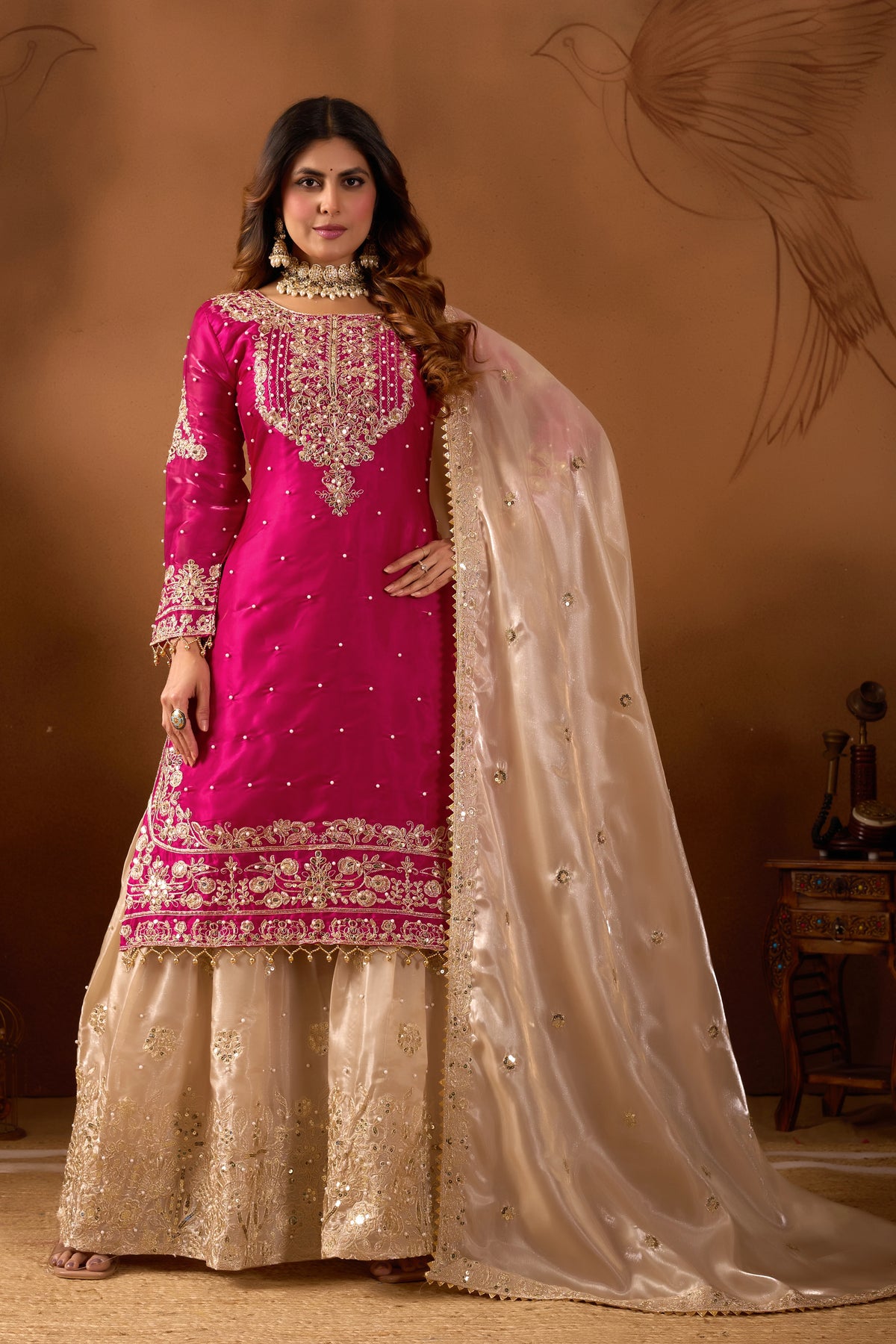 Woman wearing a pink and gold traditional outfit with a beige dupatta against a brown background