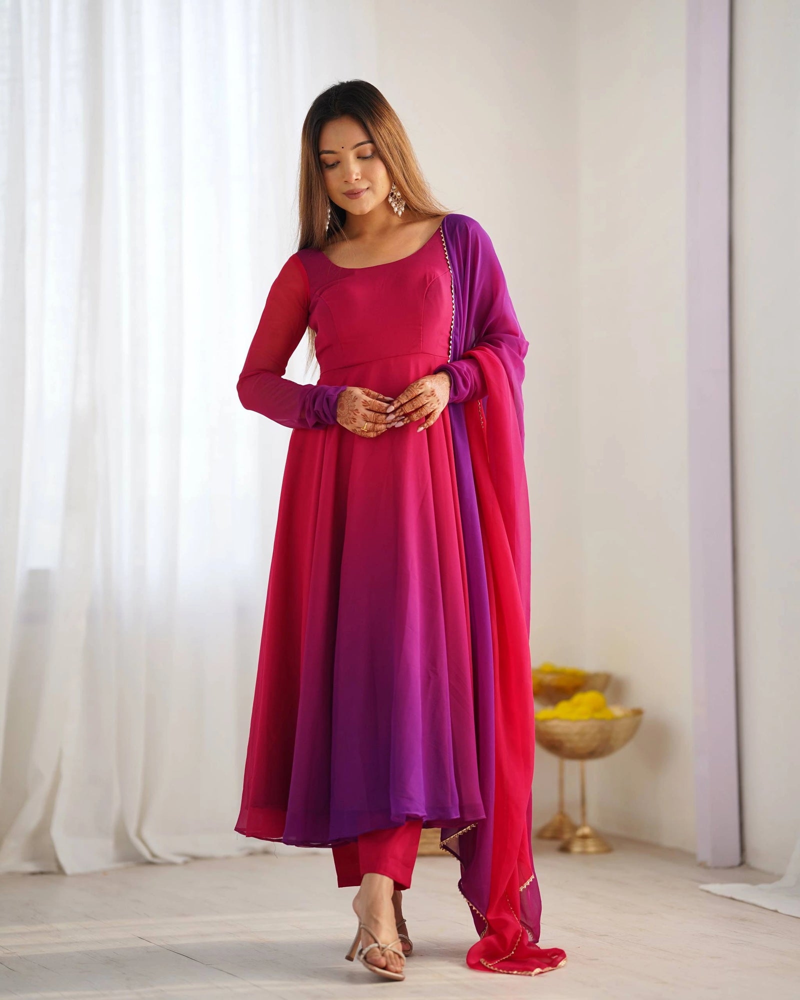 A woman stands with hands clasped, looking down, modeling a matching magenta and purple ethnic dress and pant set, with a patterned dupatta hanging around her shoulders.