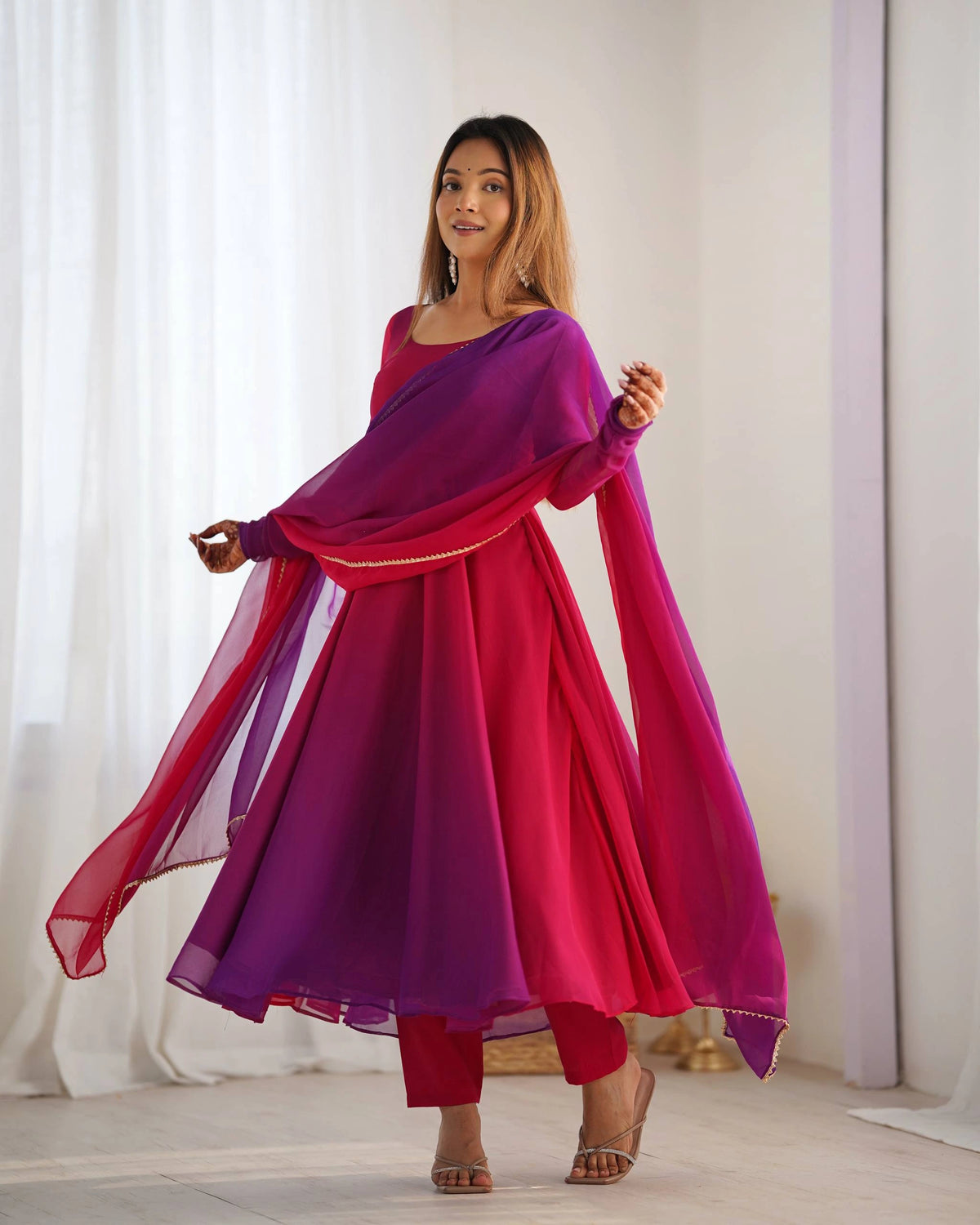 A woman modeling a long magenta and purple ethnic suit drapes her matching dupatta like a shawl, standing and smiling in a brightly lit room.