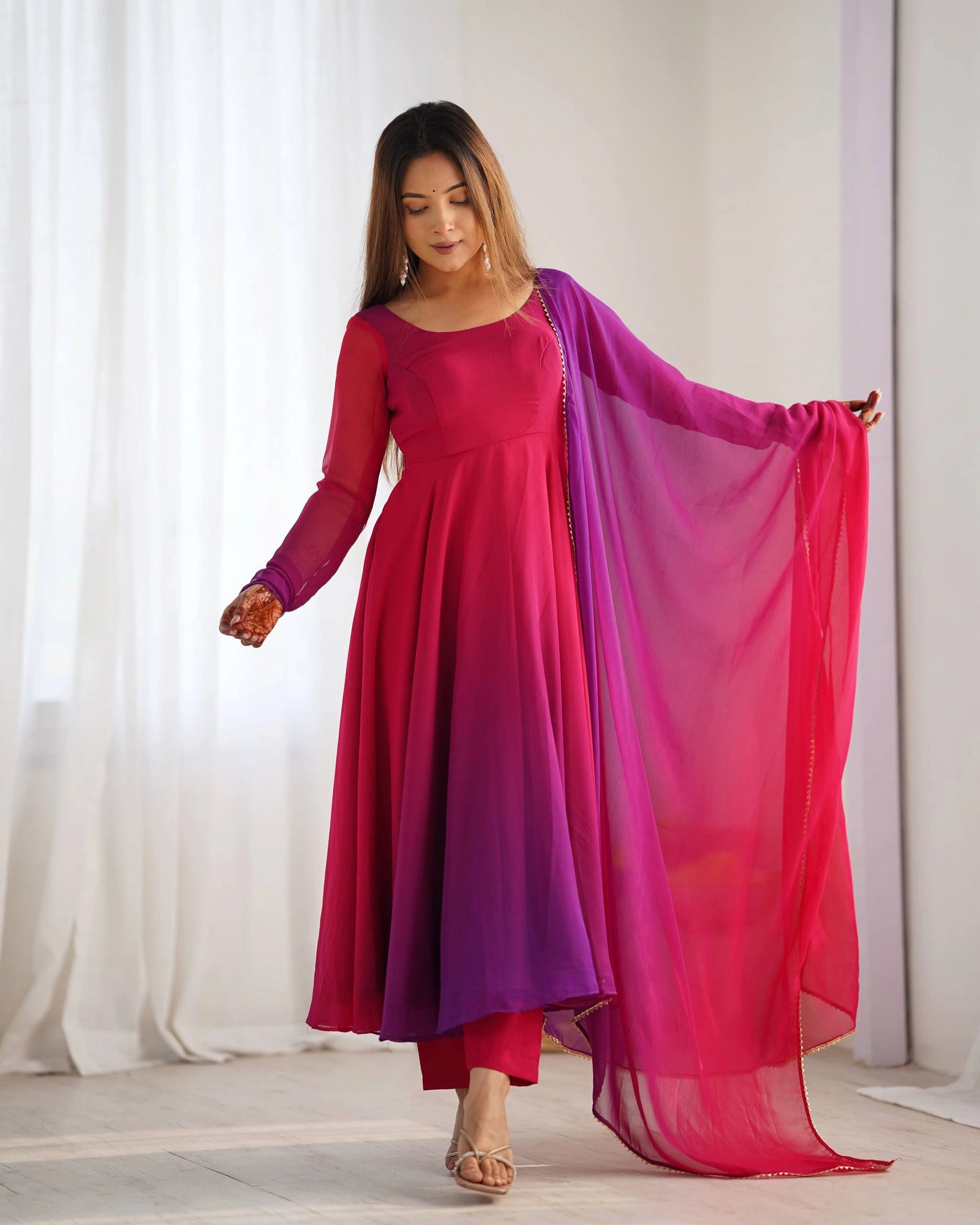 Woman wearing a pink and purple traditional outfit with a sheer pink drape against a white curtain background.