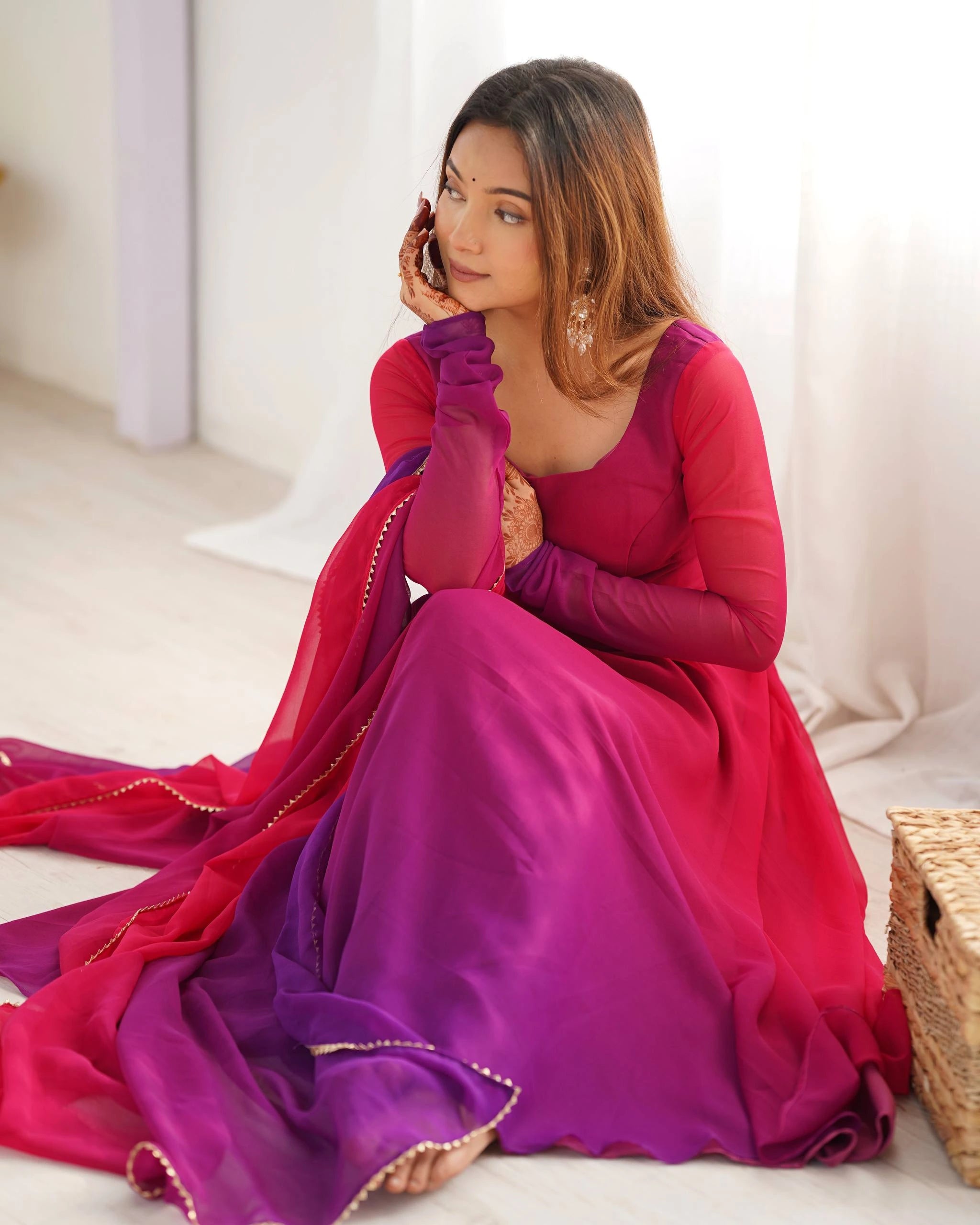 A woman sits on the floor in a vibrant magenta and purple ombre Anarkali suit with a gold-bordered dupatta. Her hands show intricate henna art as she looks away thoughtfully. She is styled with large silver drop earrings and a small bindi.