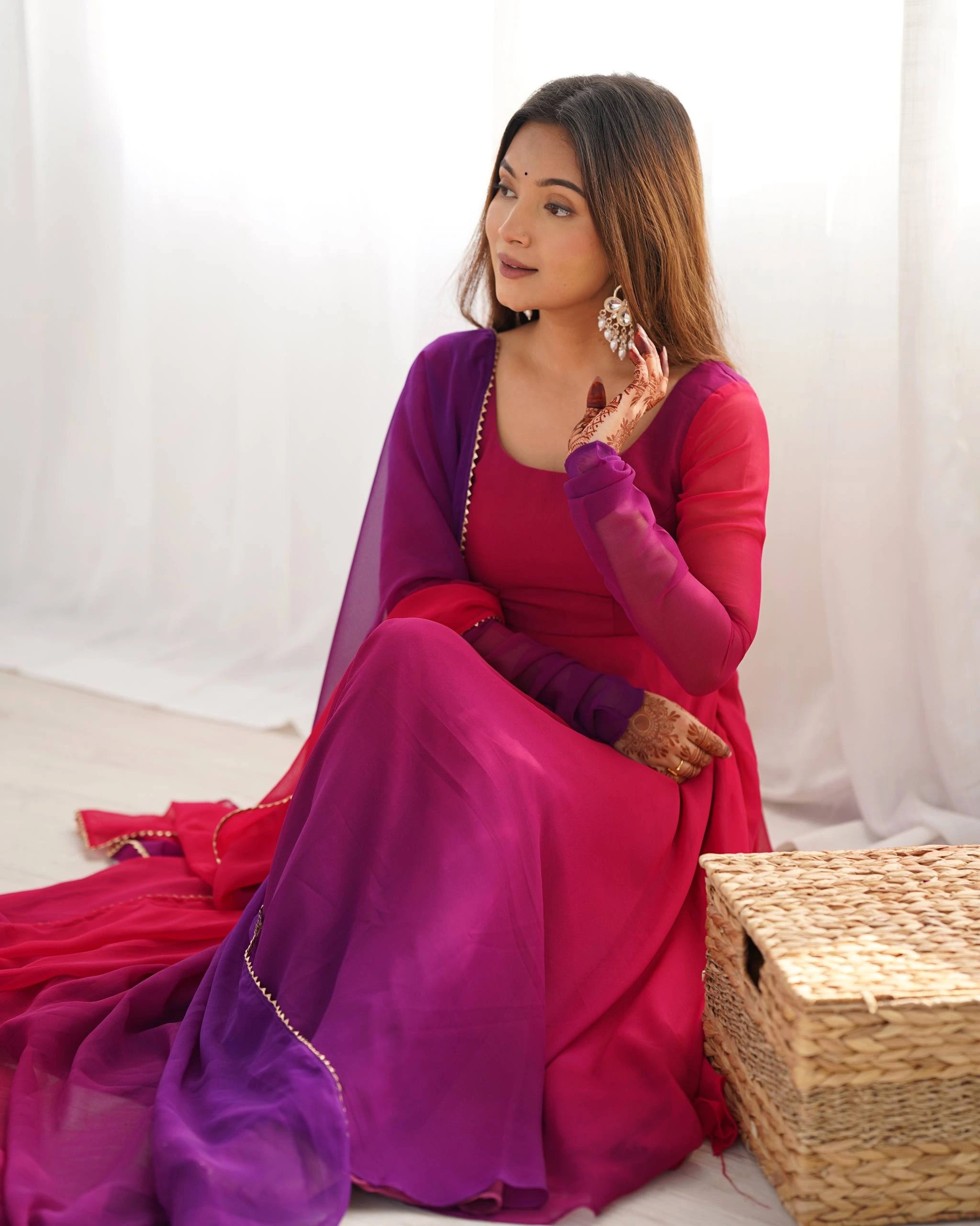 A close-up portrait of a smiling woman wearing a matching magenta and purple ethnic suit, with her hands resting on her chest showcasing detailed henna patterns.