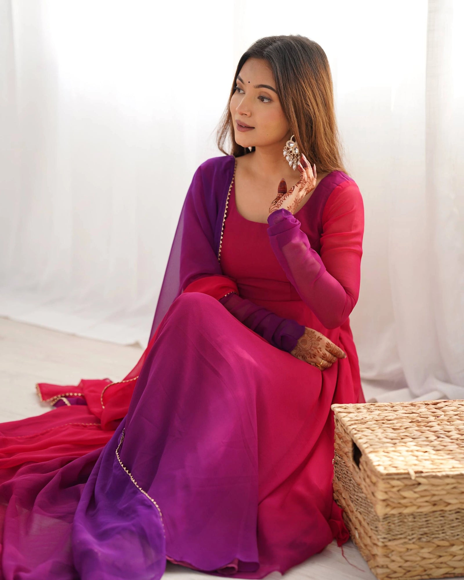 A close-up portrait of a smiling woman wearing a matching magenta and purple ethnic suit, with her hands resting on her chest showcasing detailed henna patterns.