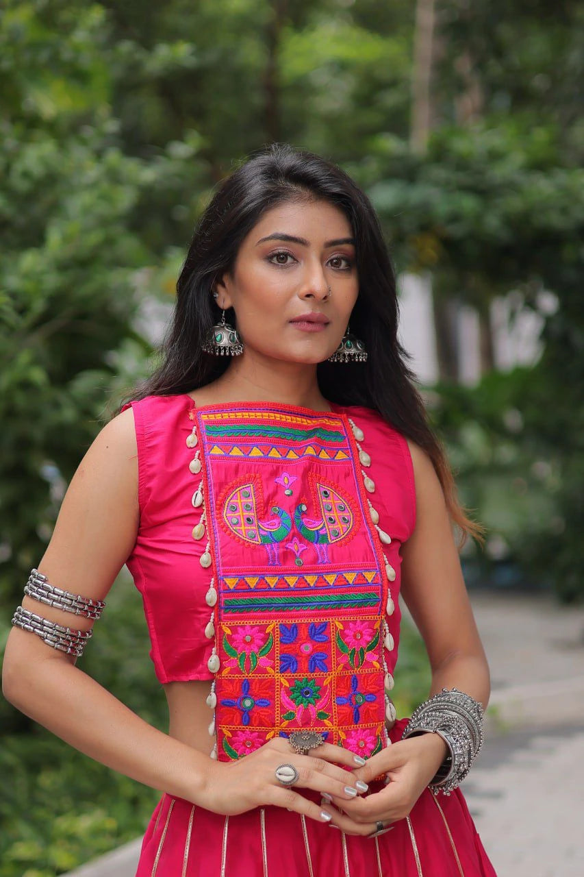 Woman wearing a vibrant traditional outfit with embroidery, standing outdoors with greenery in the background.