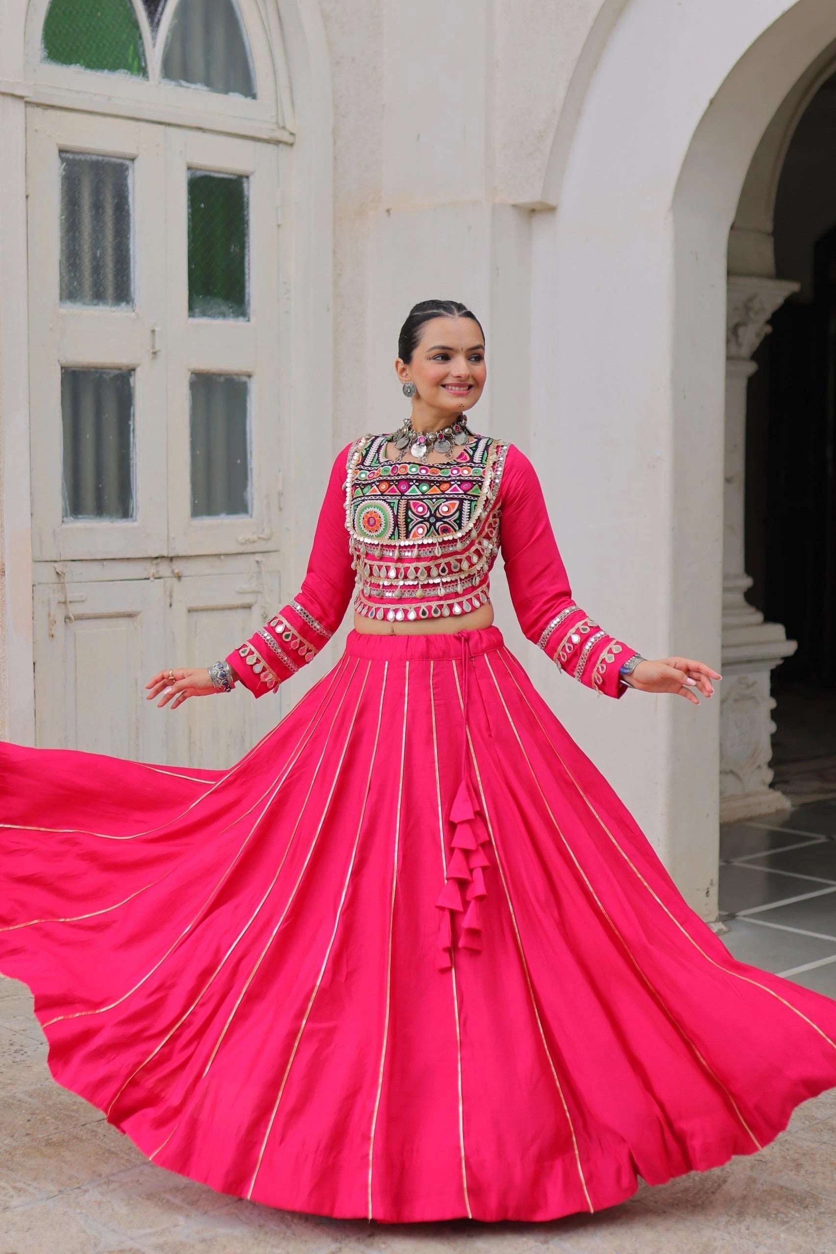 Woman in a bright pink traditional outfit with intricate patterns standing in front of a white building.
