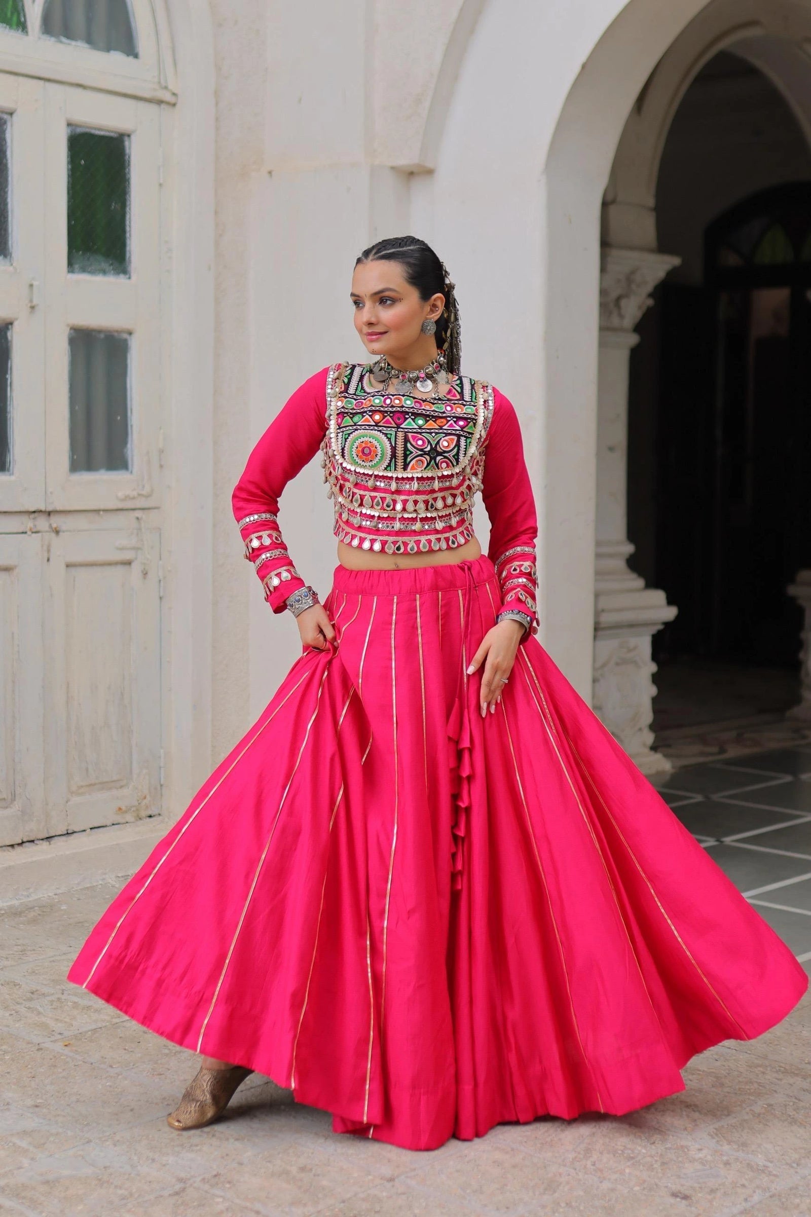 Woman in traditional pink outfit with intricate design standing in front of a white archway.