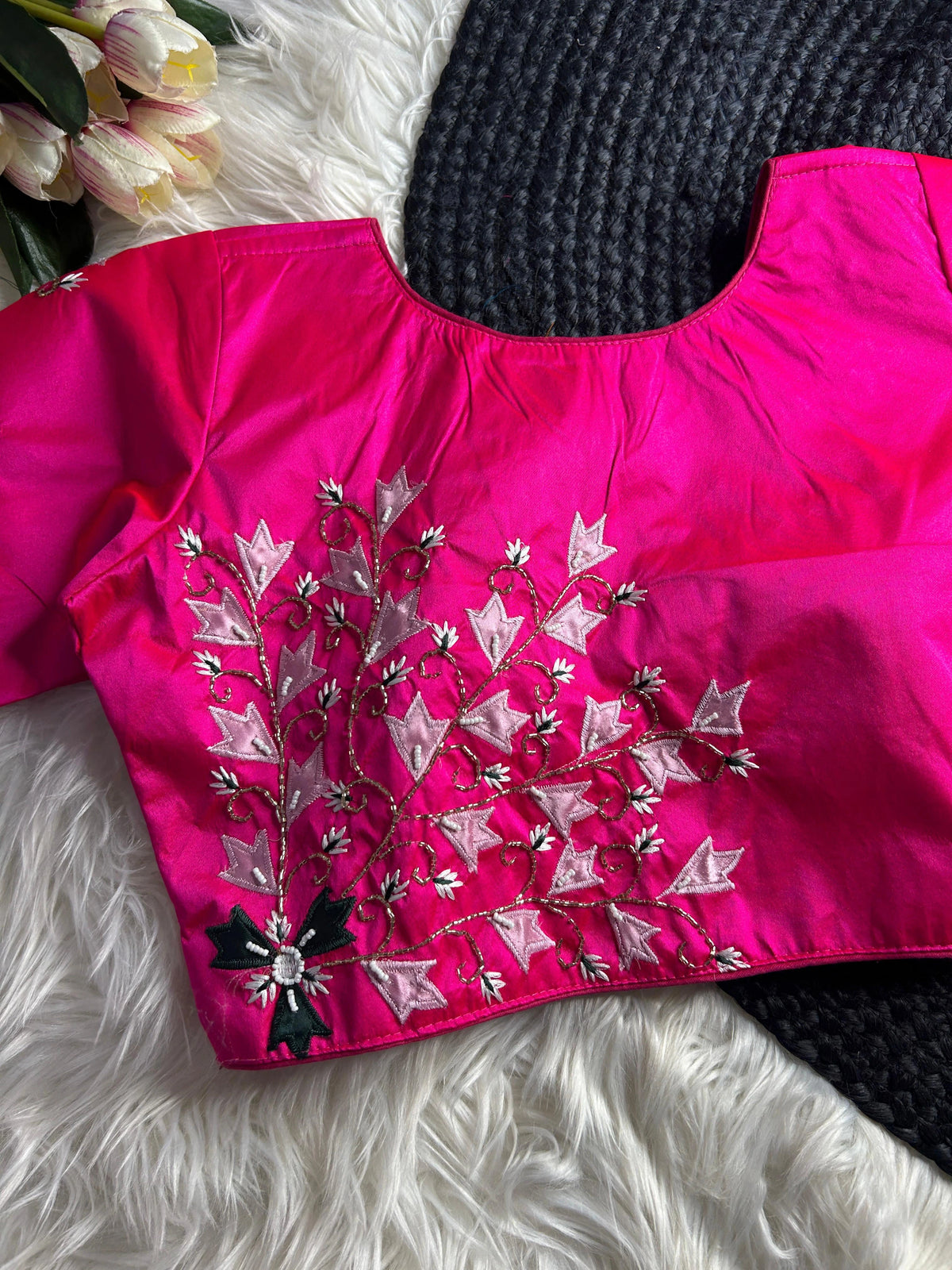 A vibrant hot pink silk blouse with a round neckline, featuring intricate hand-embroidery on the lower left front. The design consists of winding gold-beaded vines adorned with light pink silken leaves and small white floral accents, anchored by a dark green floral motif with a mirrored center at the base. The blouse is displayed flat on a white faux-fur surface alongside a gray woven mat and a cluster of pink and white tulips.