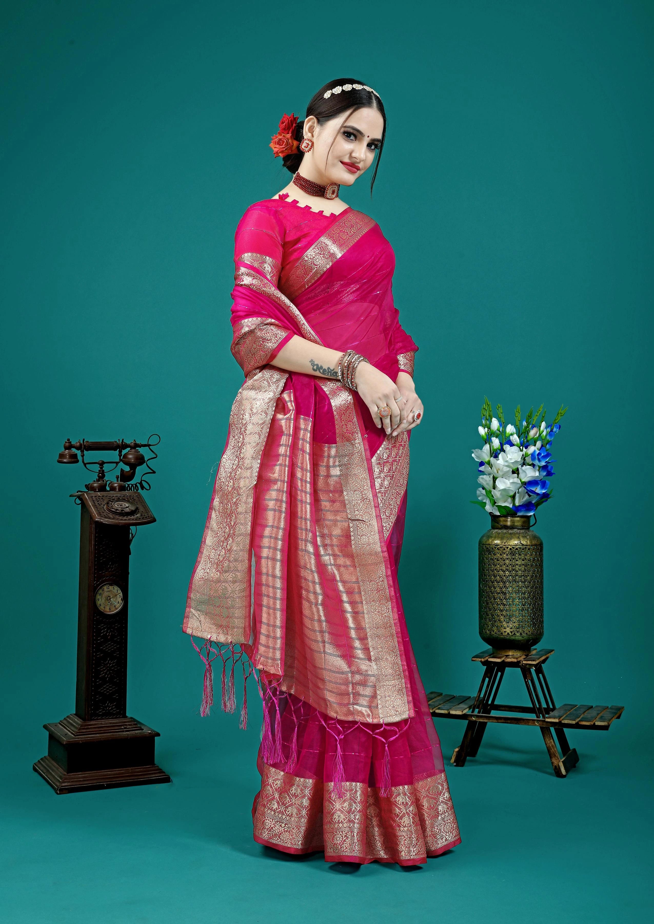 Woman in a pink saree standing against a teal background with decorative elements.