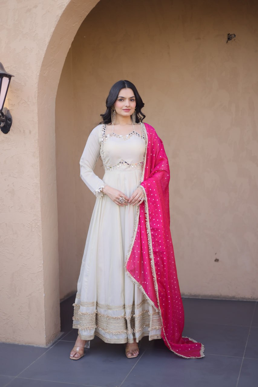 Woman in a white traditional outfit with a pink dupatta standing in front of a beige wall.